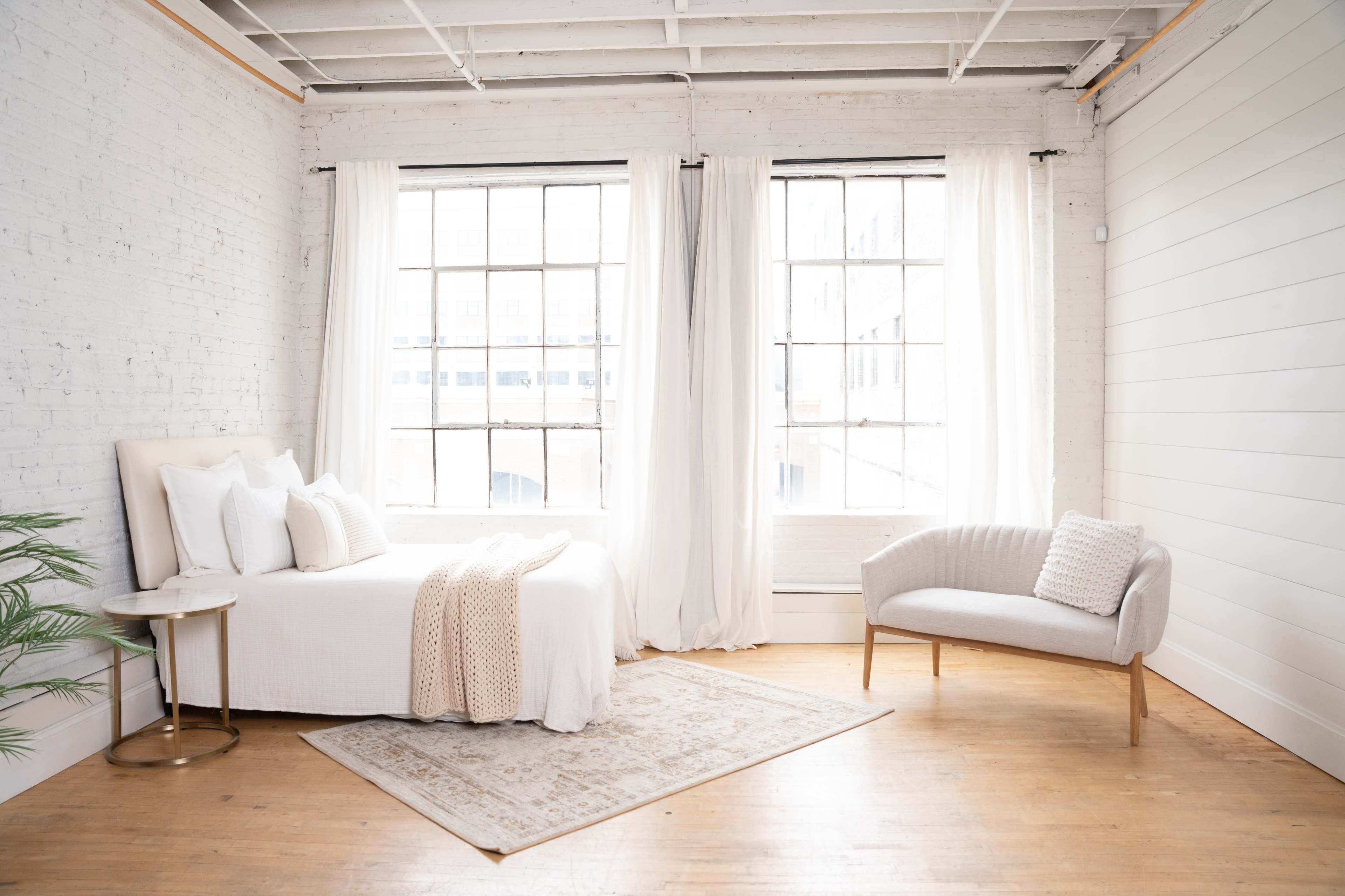 The image shows a minimalist bedroom featuring a bed with white linens, large windows with sheer curtains, and a light-colored armchair beside a small plant.