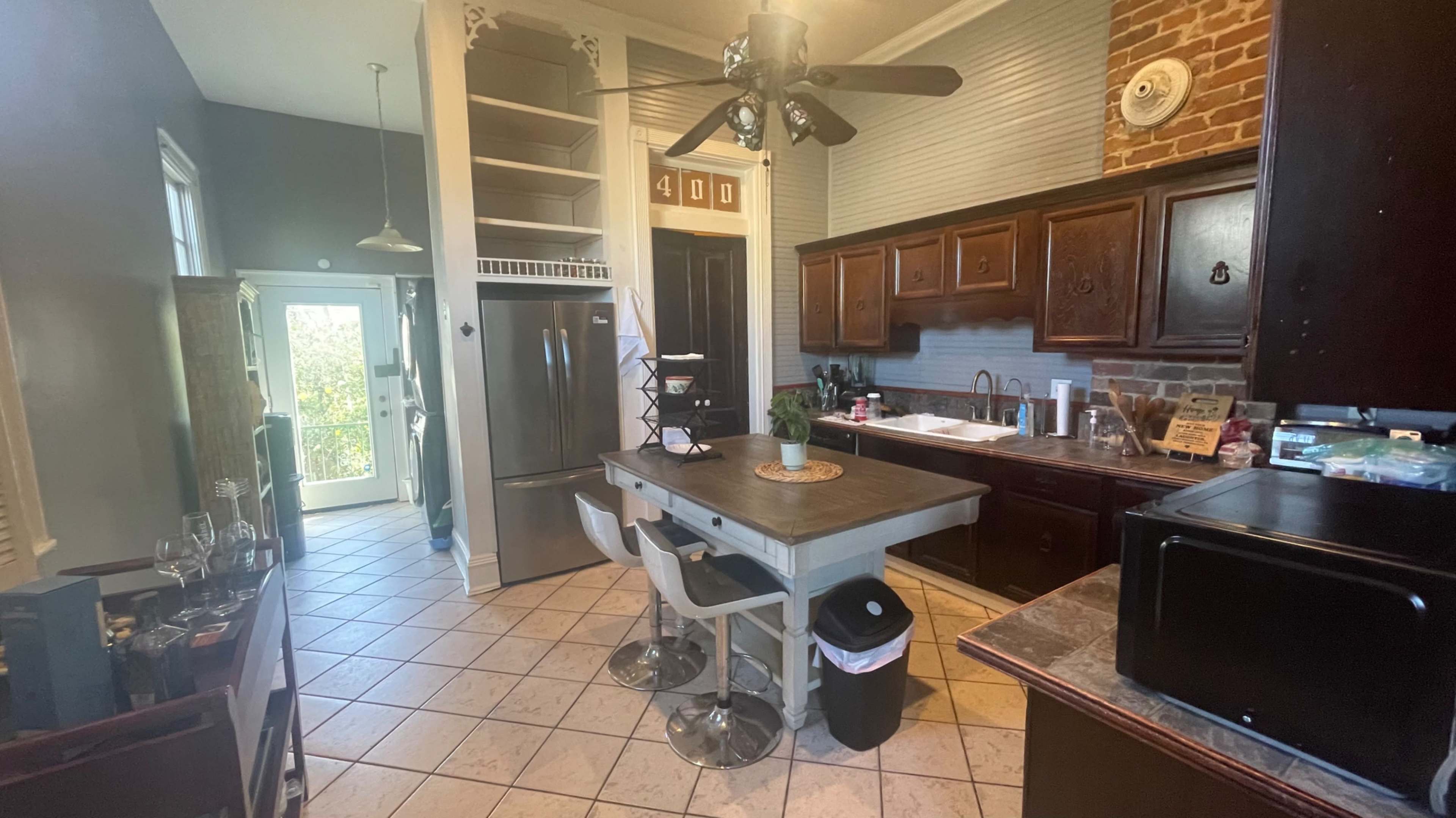 The image shows a kitchen with a mix of brick and wooden cabinetry, a central table with stools, and a doorway leading to another room.