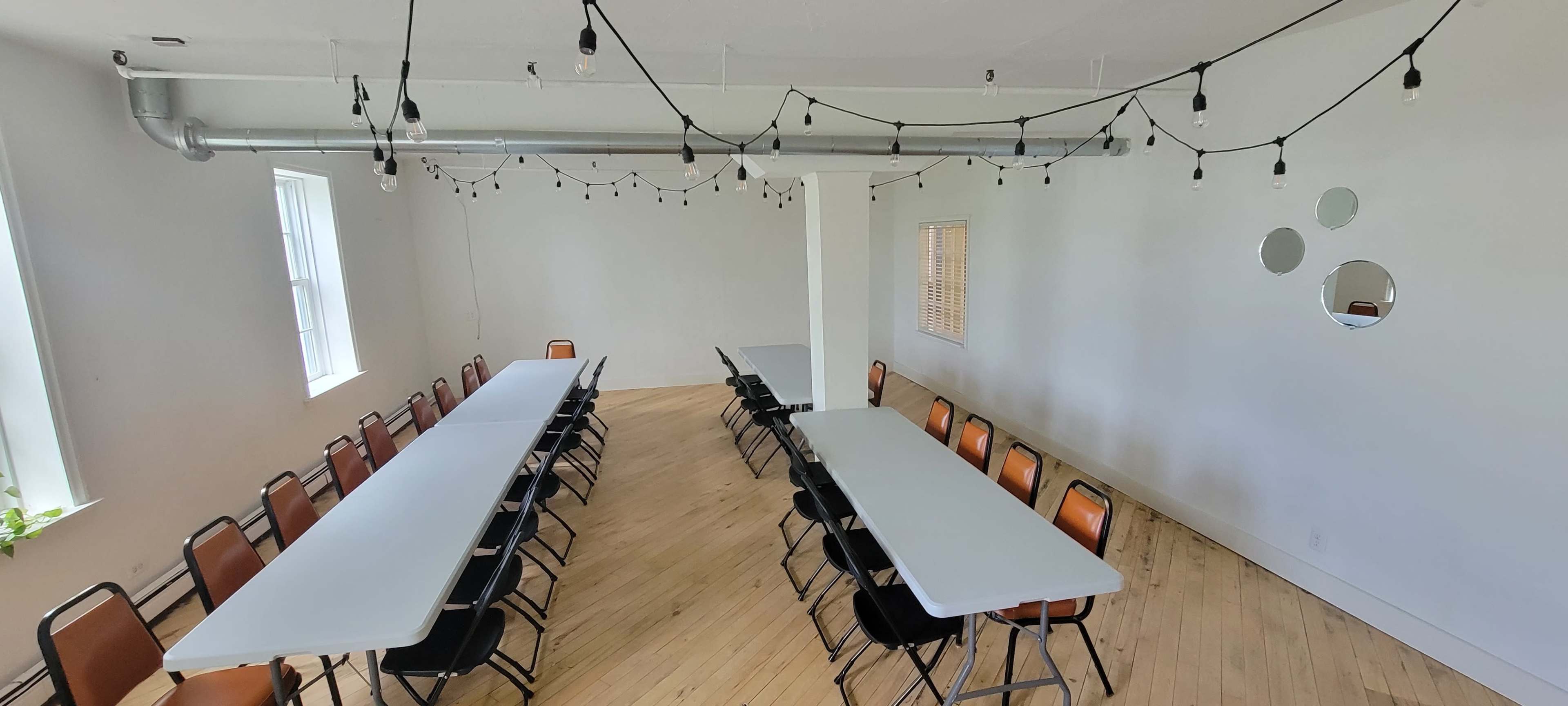 A spacious, minimalist room features two long tables arranged in a U-shape, surrounded by chairs and illuminated by string lights overhead.