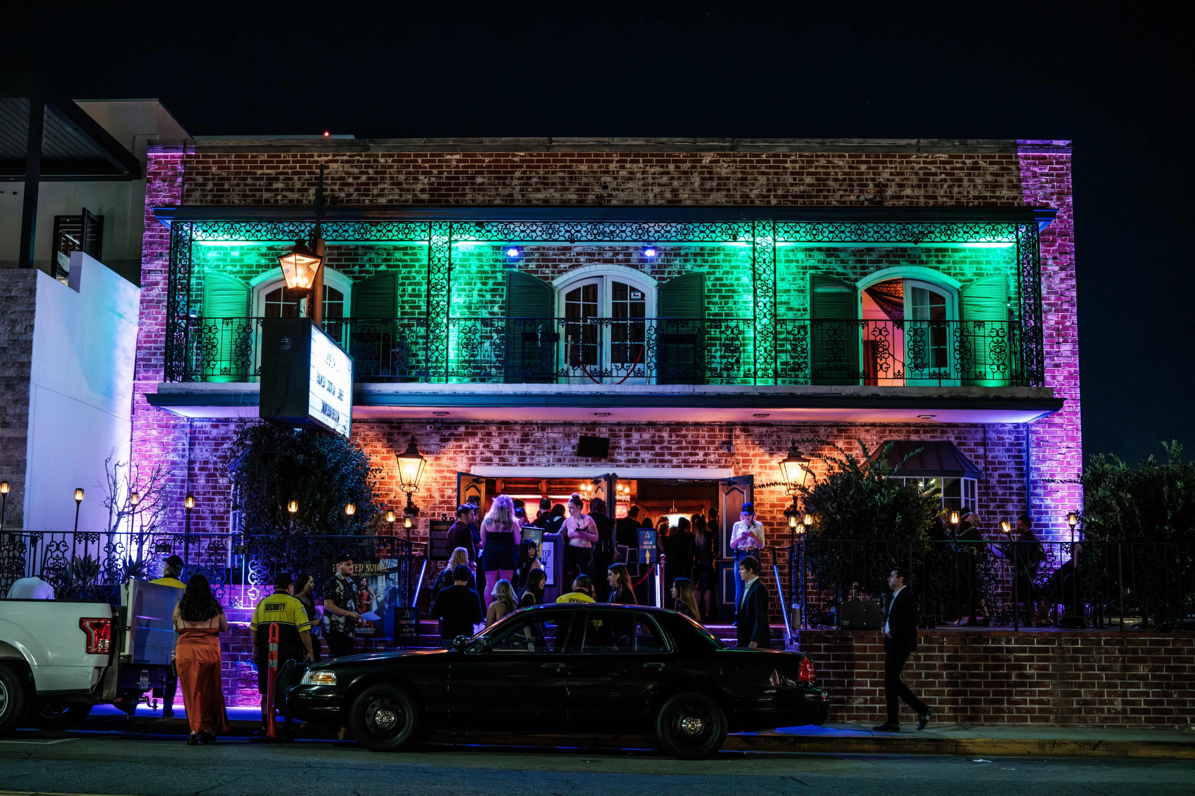 A lively gathering of people takes place outside a brightly lit, two-story brick building at night.