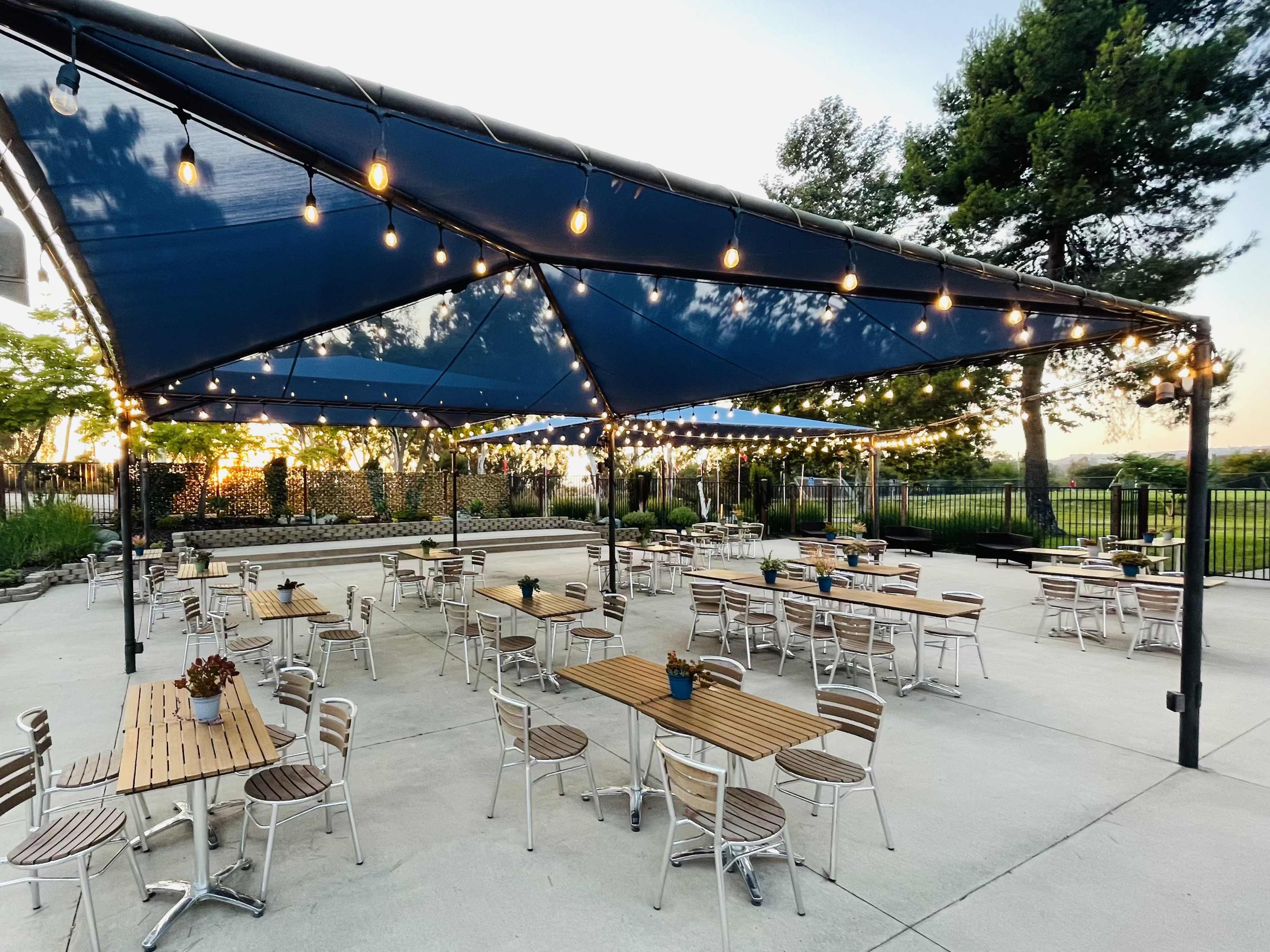 An outdoor dining area features multiple wooden tables and chairs under a large blue canopy with string lights, surrounded by greenery.