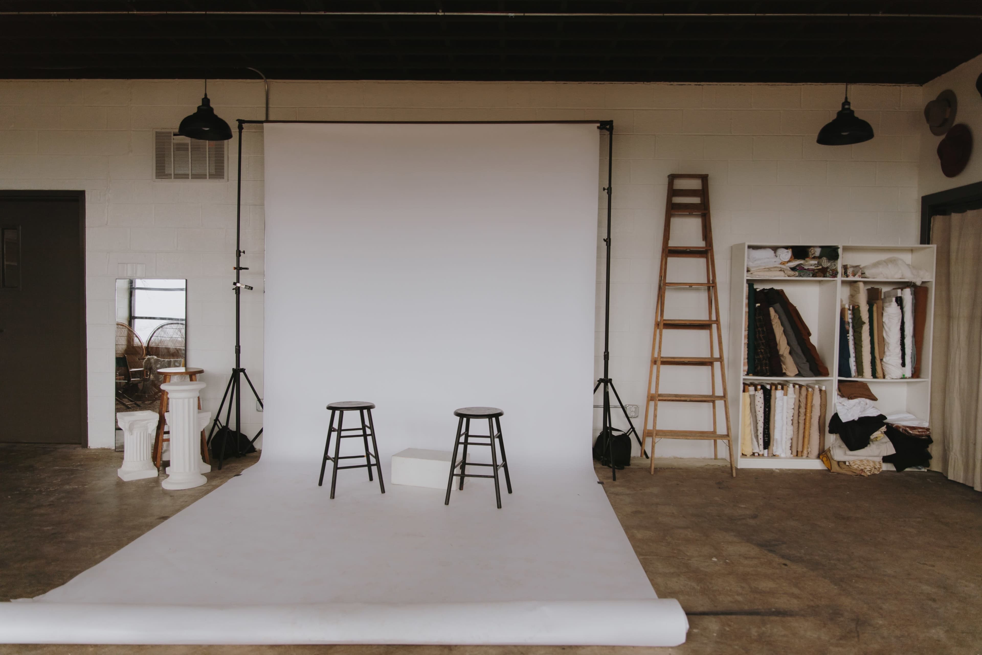 The image shows a photography studio with a white backdrop, two black stools, a ladder, and a shelving unit filled with fabric rolls.