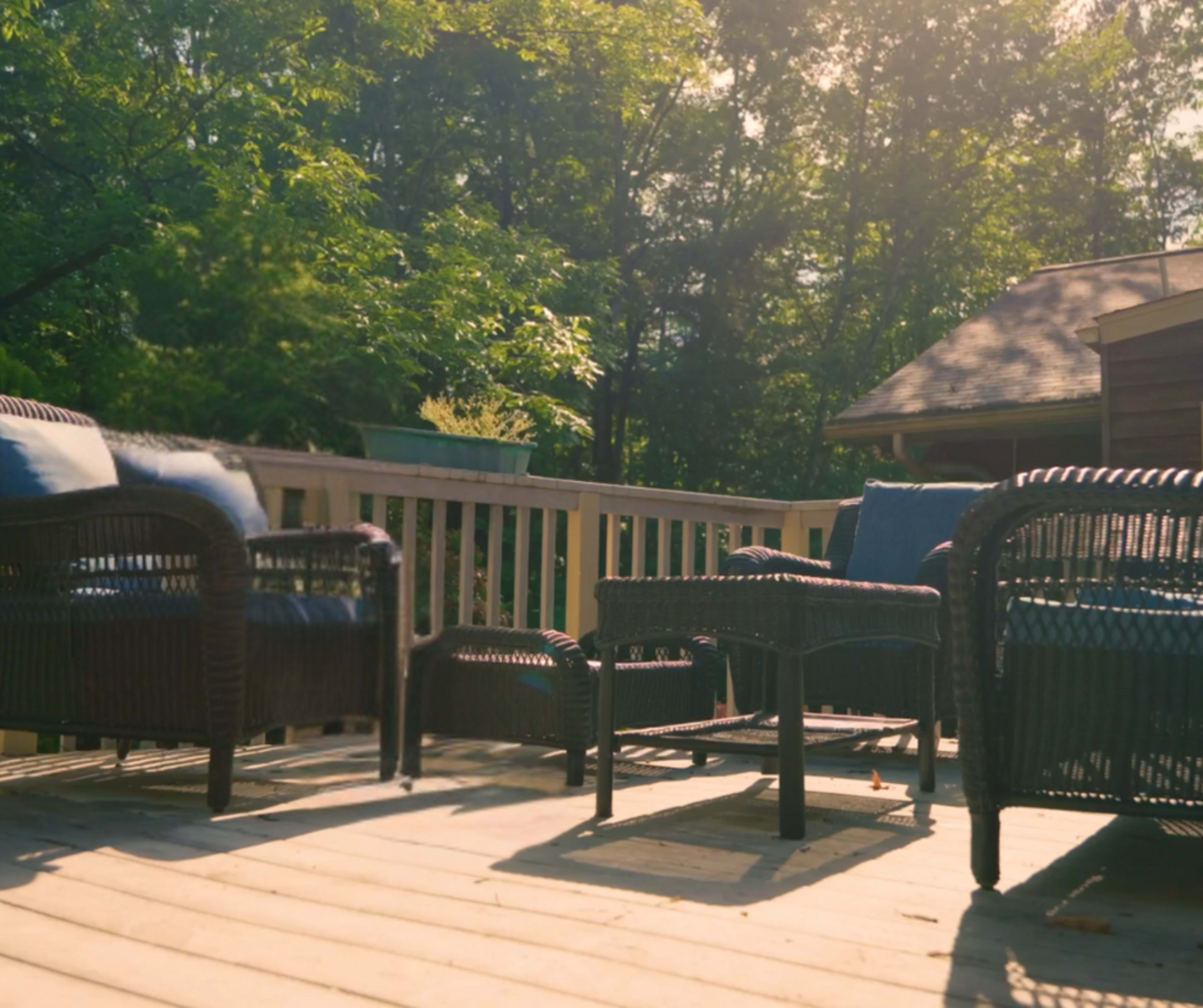 The image shows a set of woven patio furniture arranged on a wooden deck surrounded by trees.