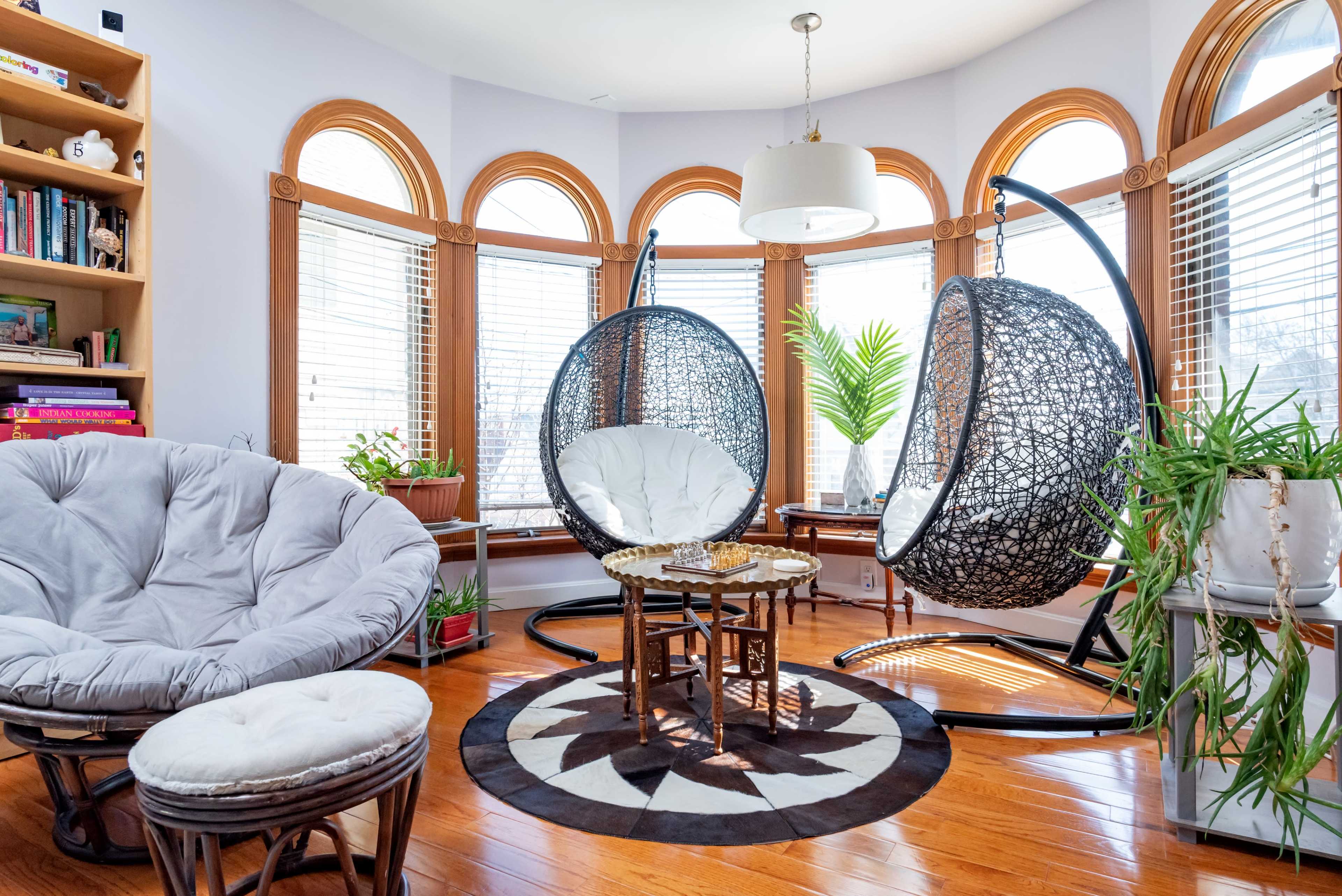 A sunlit room features two black hanging chairs, a round table, and a plush seated chair, surrounded by large windows and indoor plants.