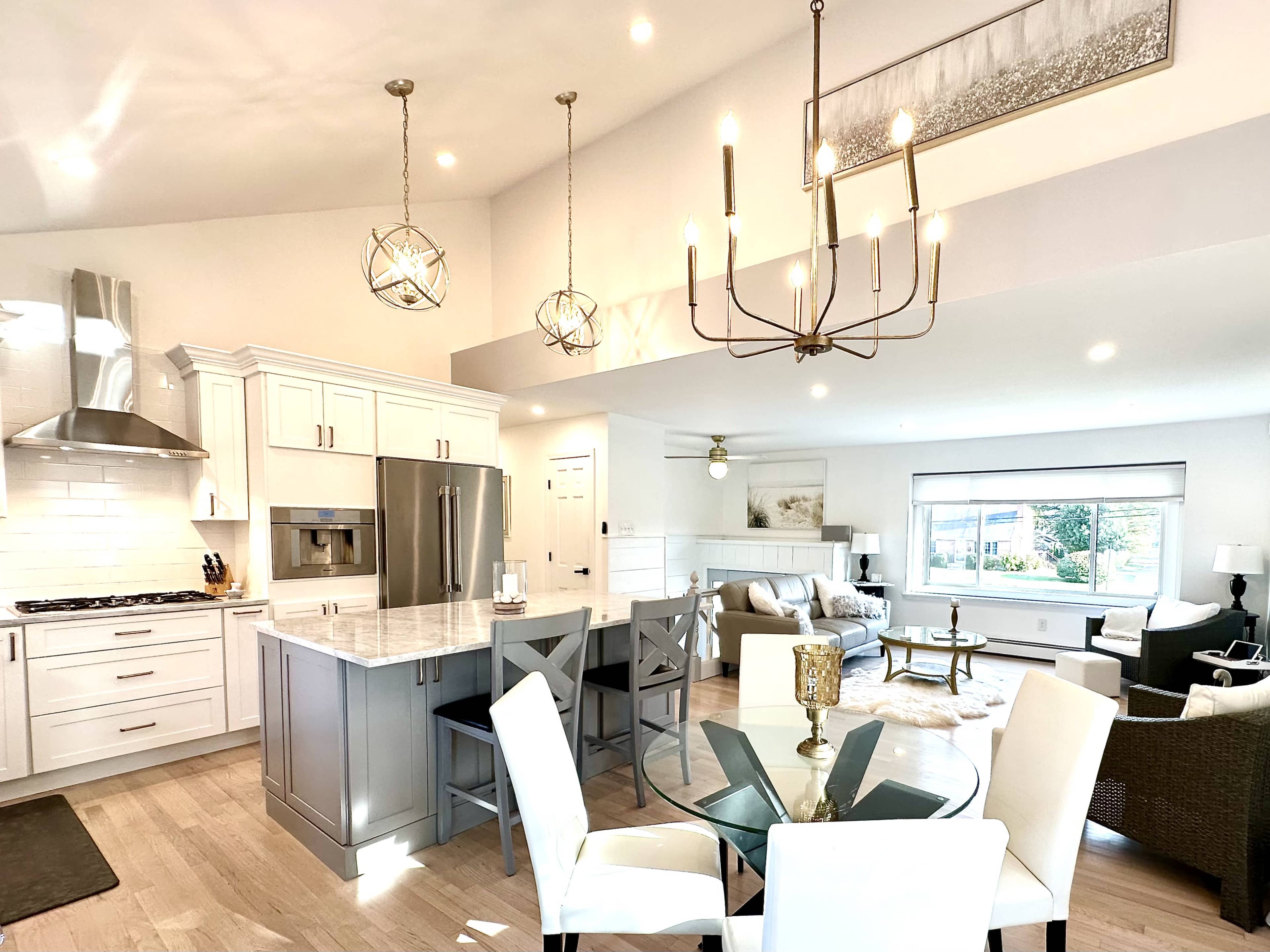 The image depicts a modern open-concept kitchen and living area featuring white cabinetry, a large island with seating, and a seating area with a sofa and armchairs.