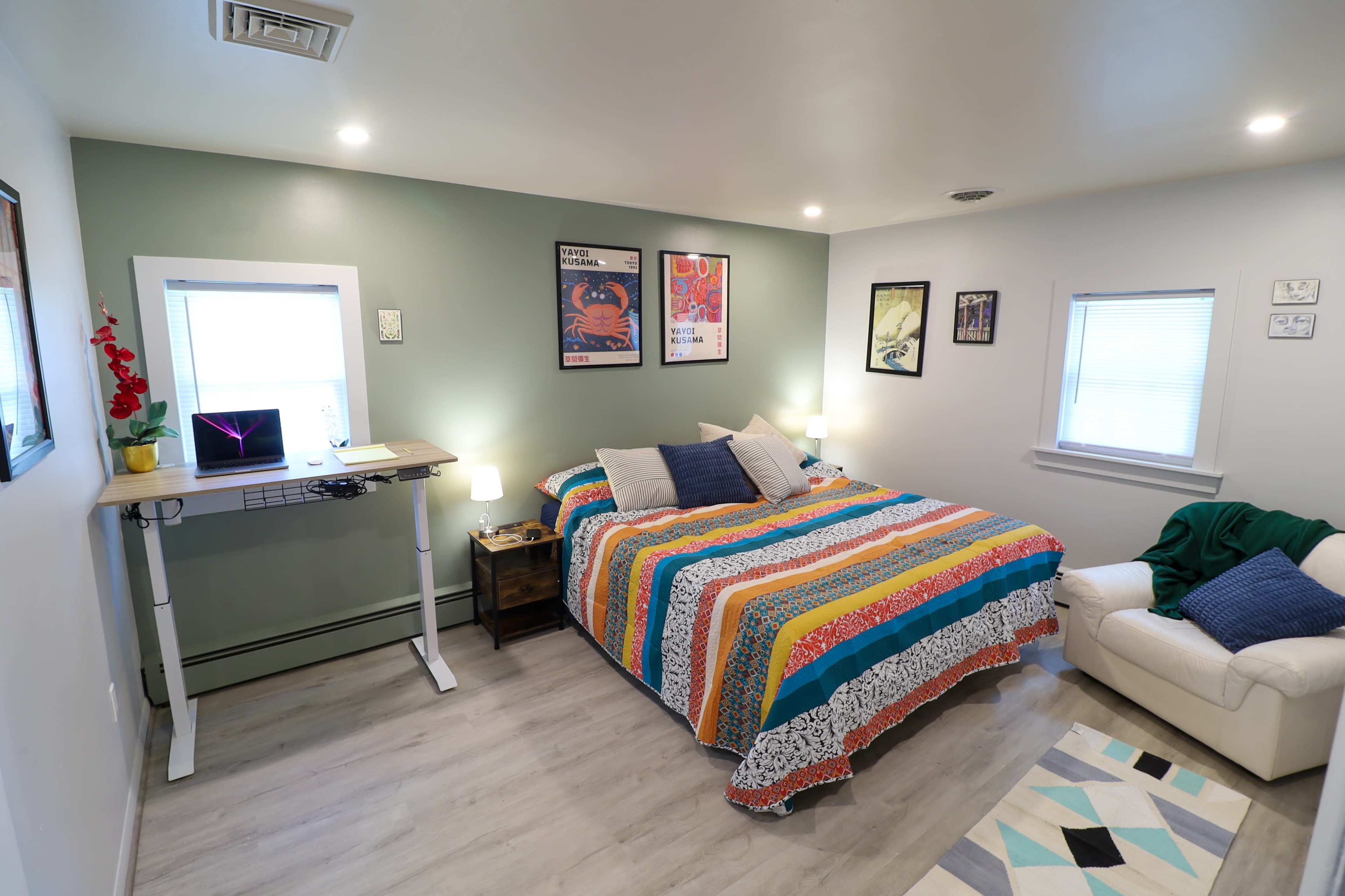 A well-lit bedroom featuring a colorful patterned bedspread, a standing desk with a laptop, and a cozy armchair.