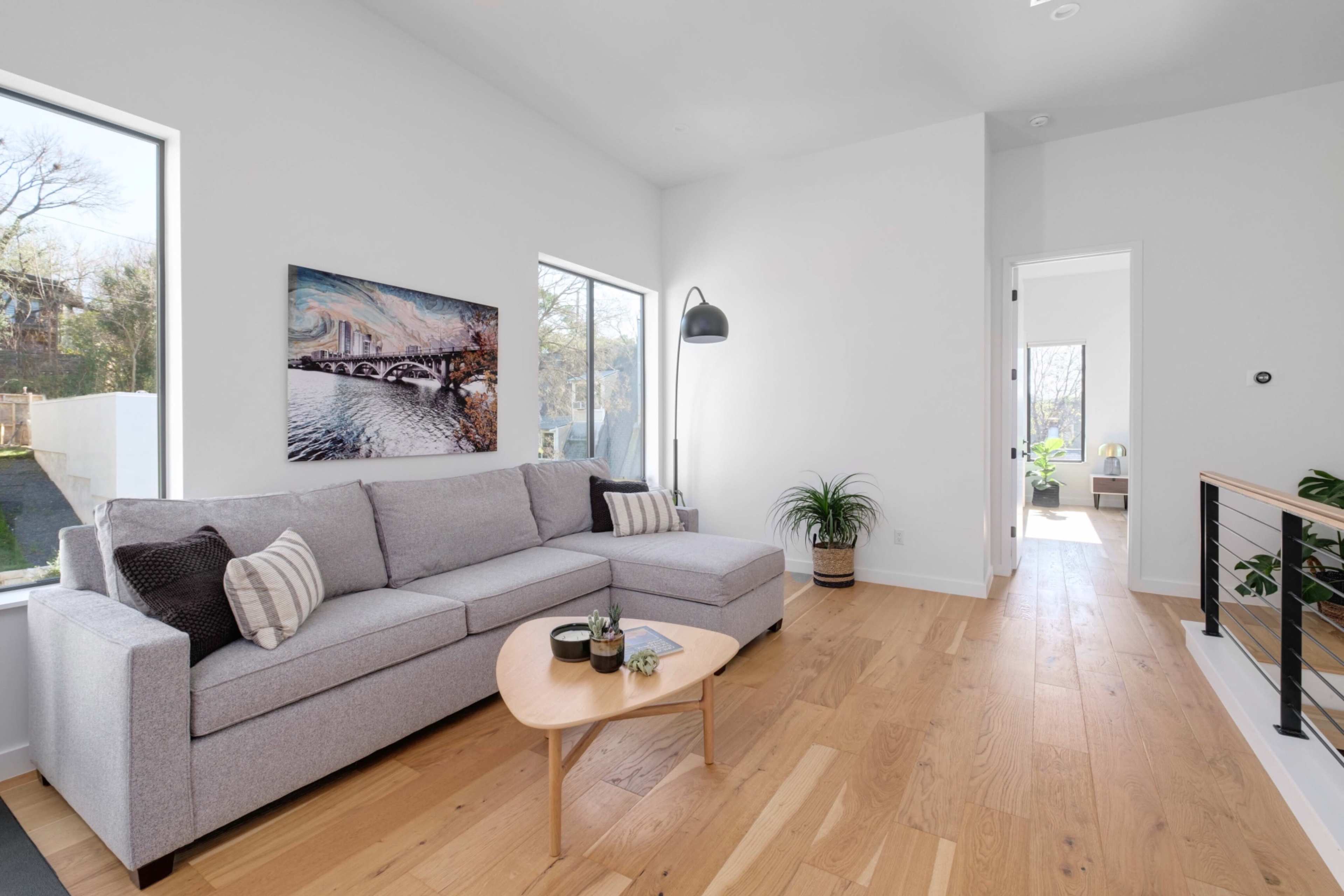 A light-filled living room features a gray sofa, a wooden coffee table, and a painting of a bridge on the wall, with natural light illuminating the space through two large windows.