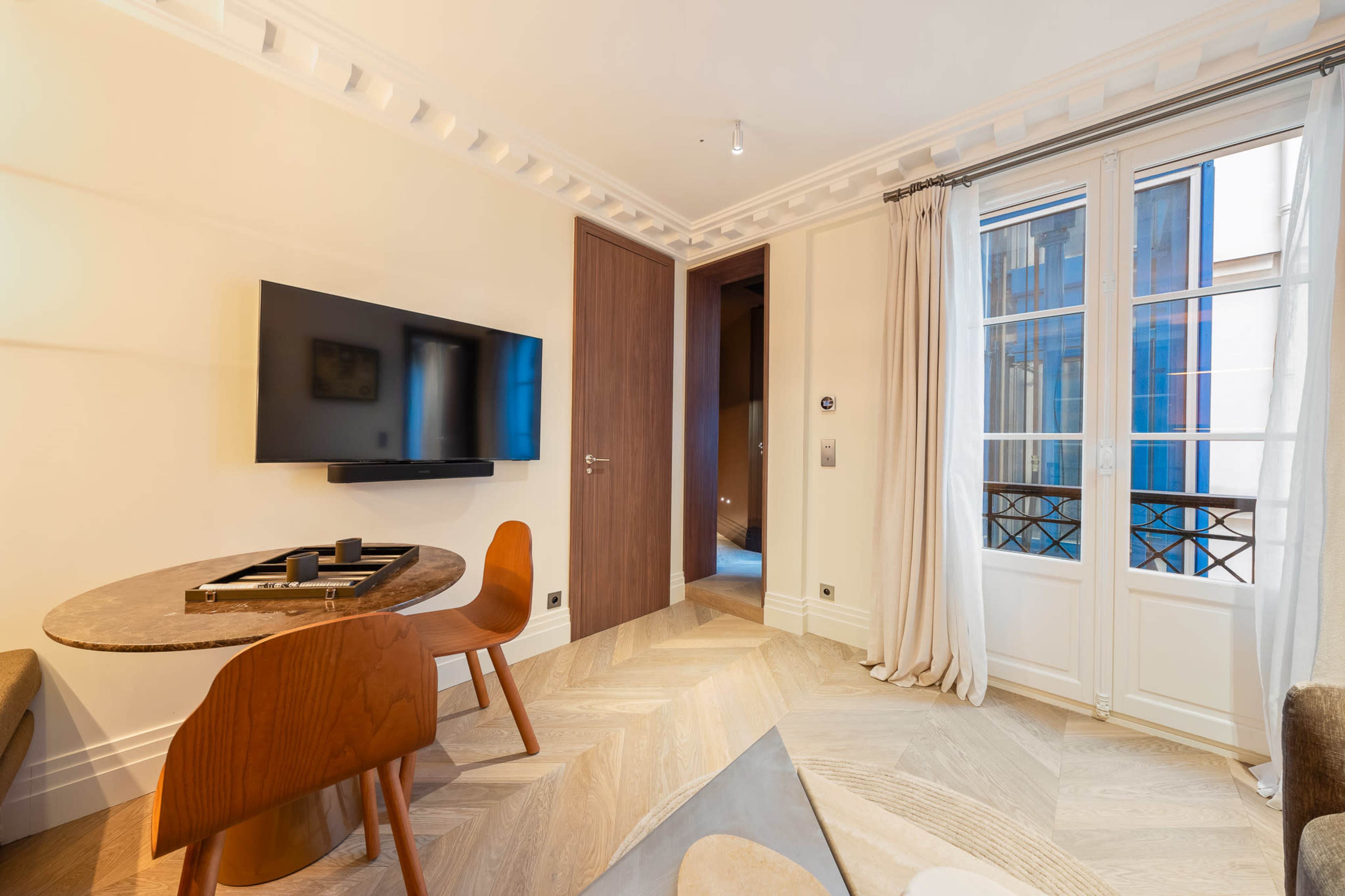 The image shows a modern living space featuring a round table with wooden chairs, a wall-mounted television, and large windows with sheer curtains.