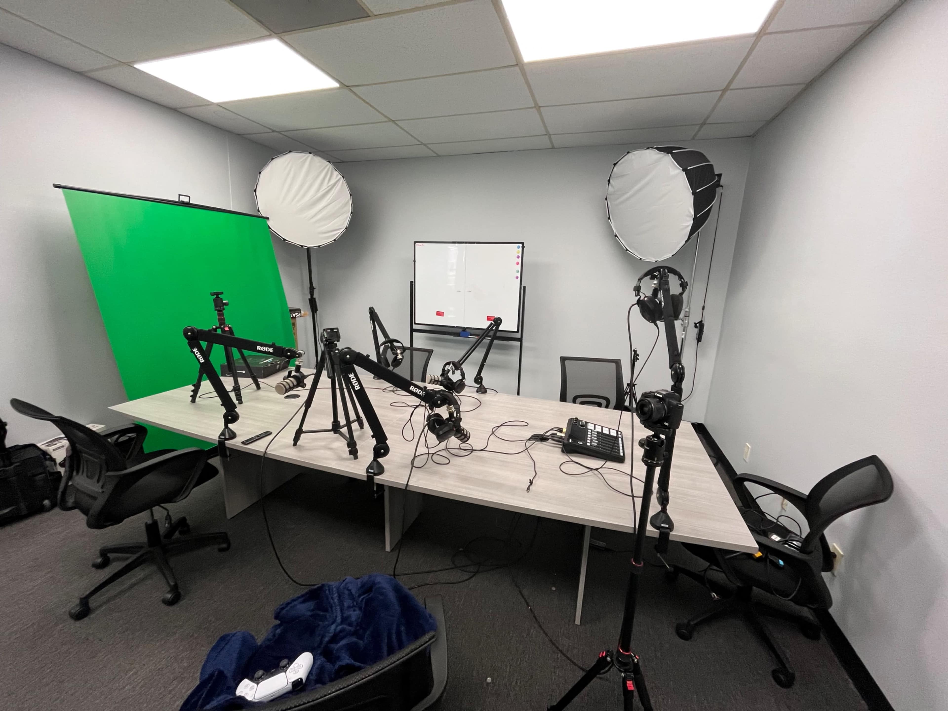 A conference room is set up with multiple camera tripods, softbox lights, a green screen, and a whiteboard.