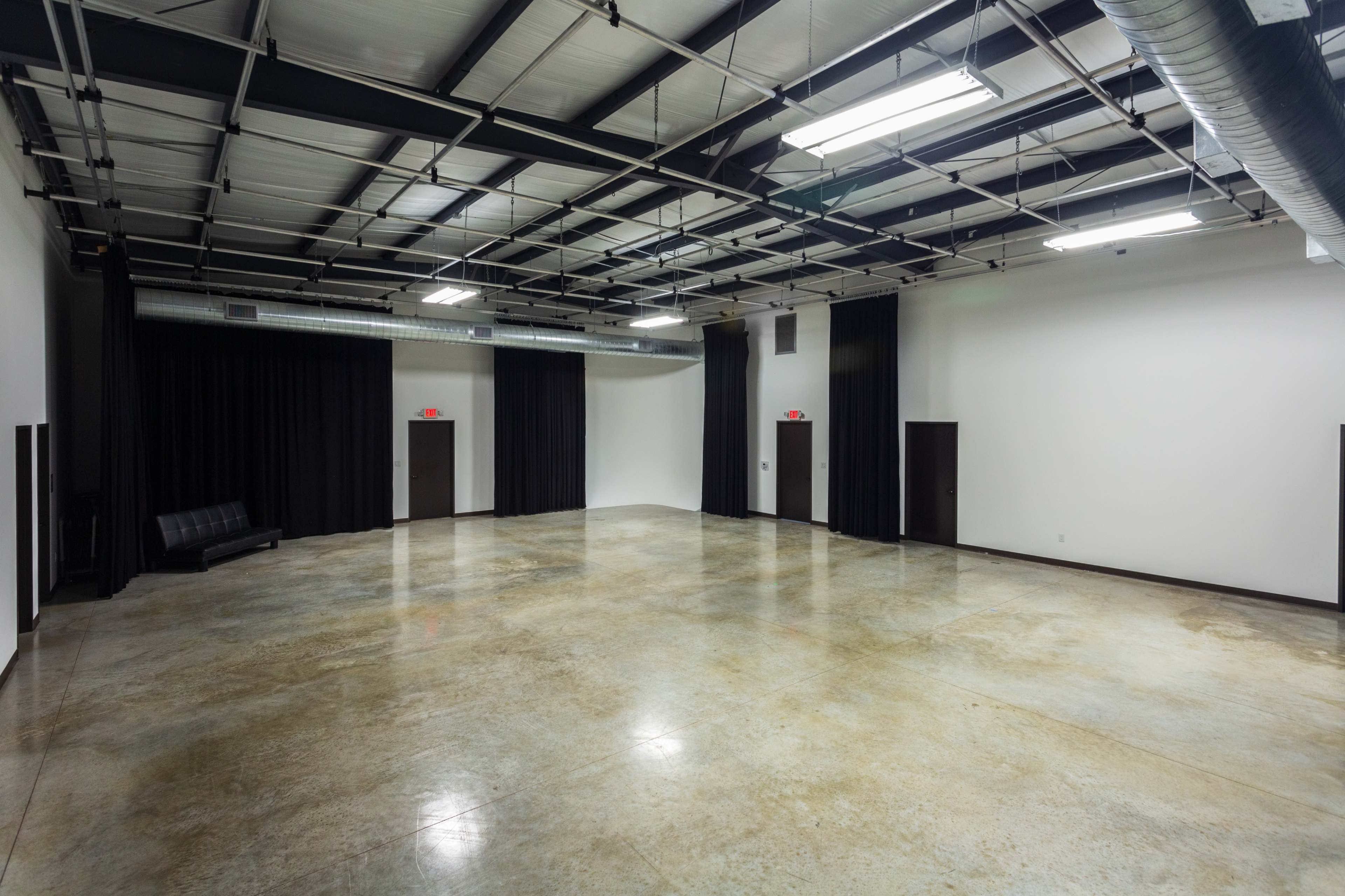 The image shows a large, empty room with concrete flooring, black curtains along the walls, and two doors on opposite sides.