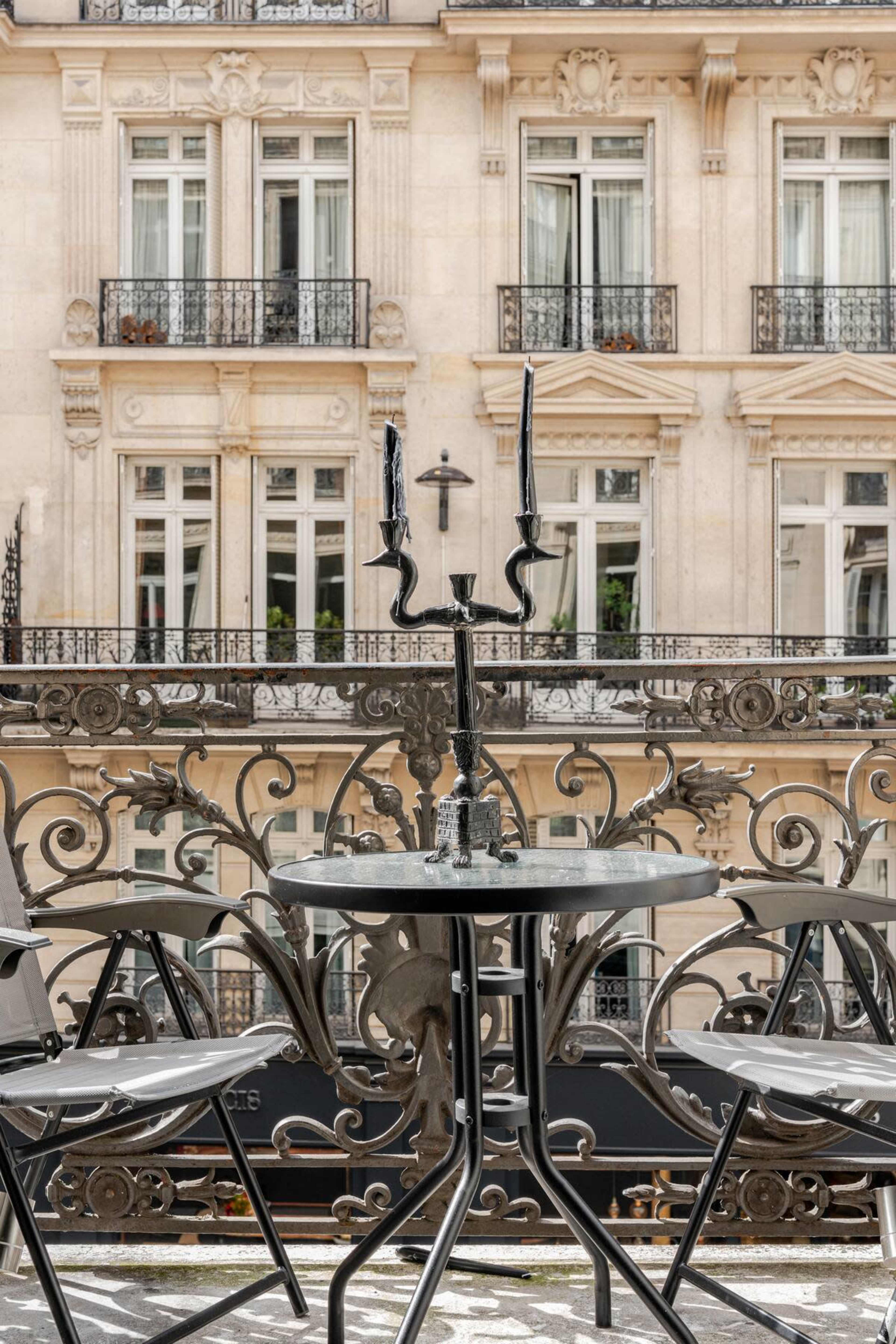 A black wrought-iron table with two chairs and a candlestick stands on a balcony overlooking a building with large windows and decorative metal railings.