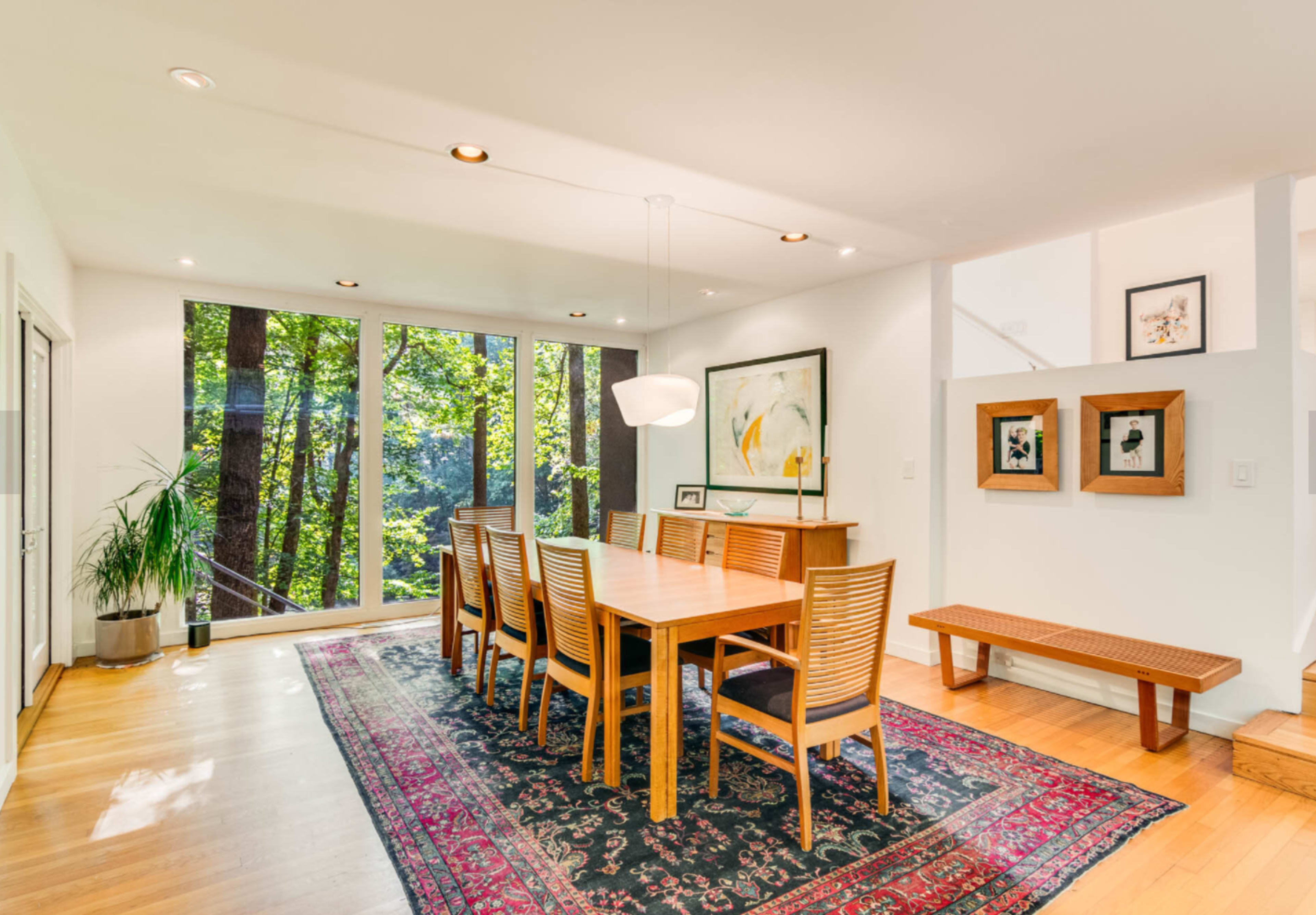 A wooden dining table with chairs is positioned in a bright room featuring large windows that open to a view of trees outside.