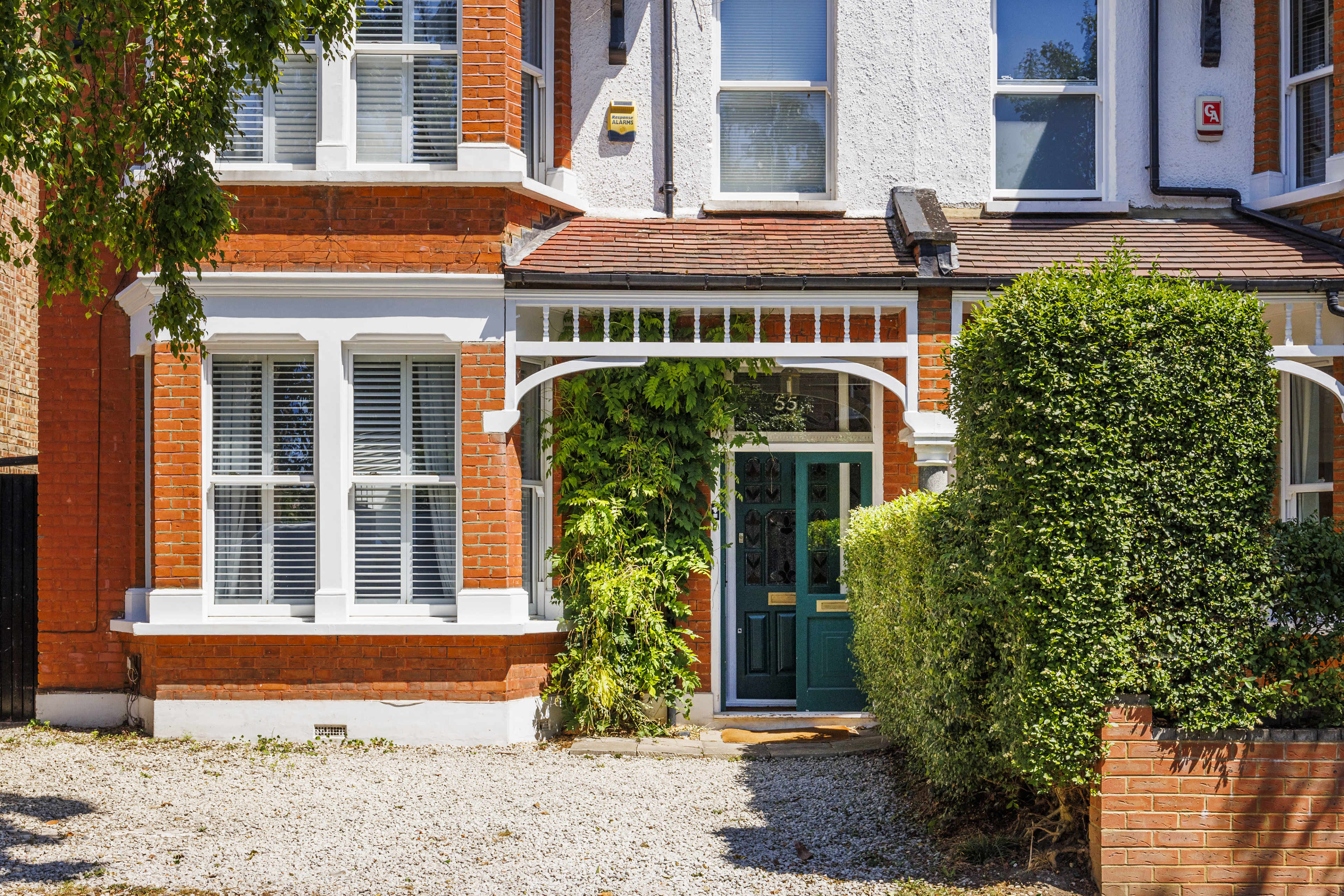 A two-story brick house features a covered entryway with green doors and surrounding greenery.