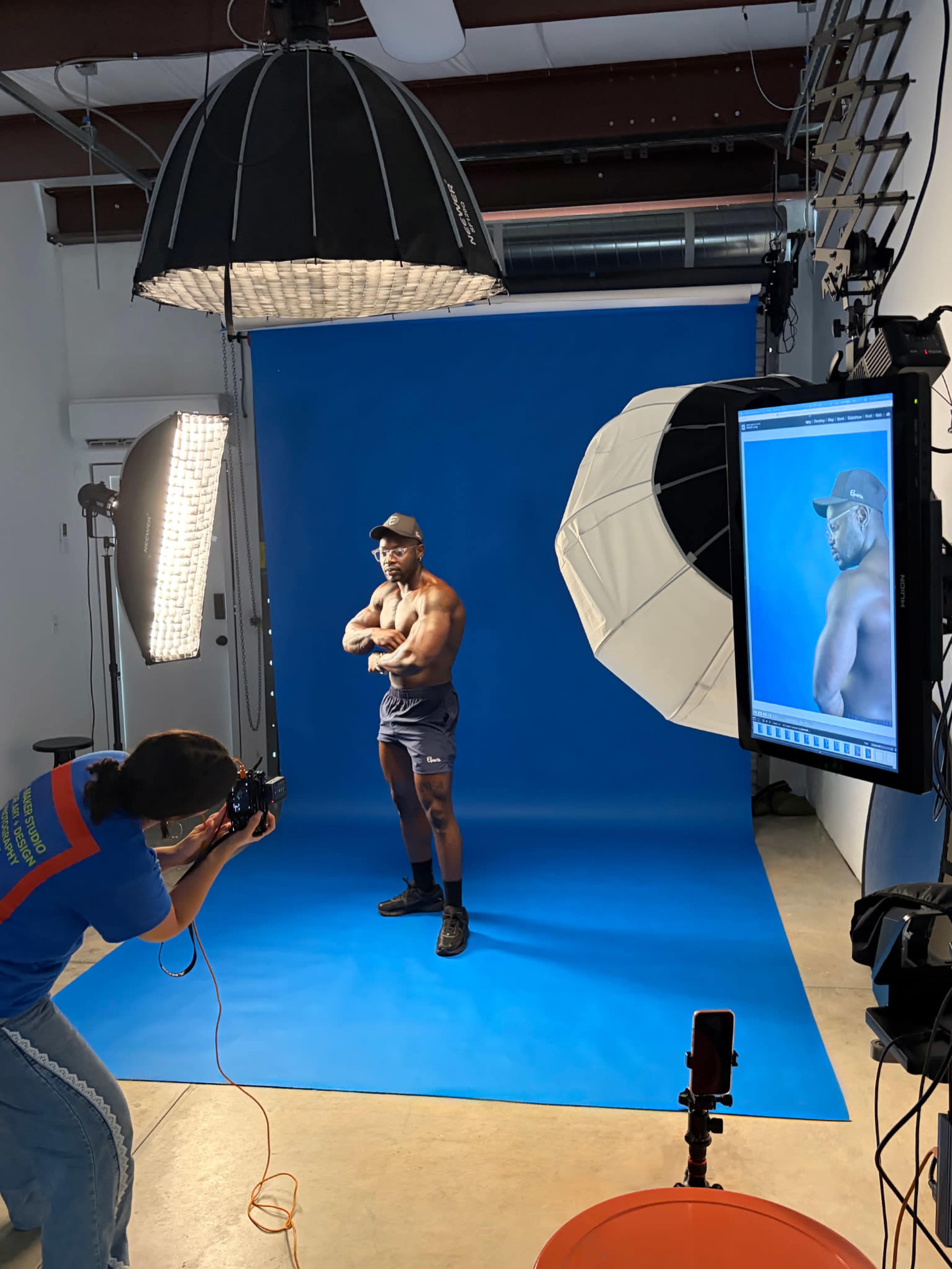 A photographer captures a muscular model posing with crossed arms in front of a bright blue backdrop in a studio.