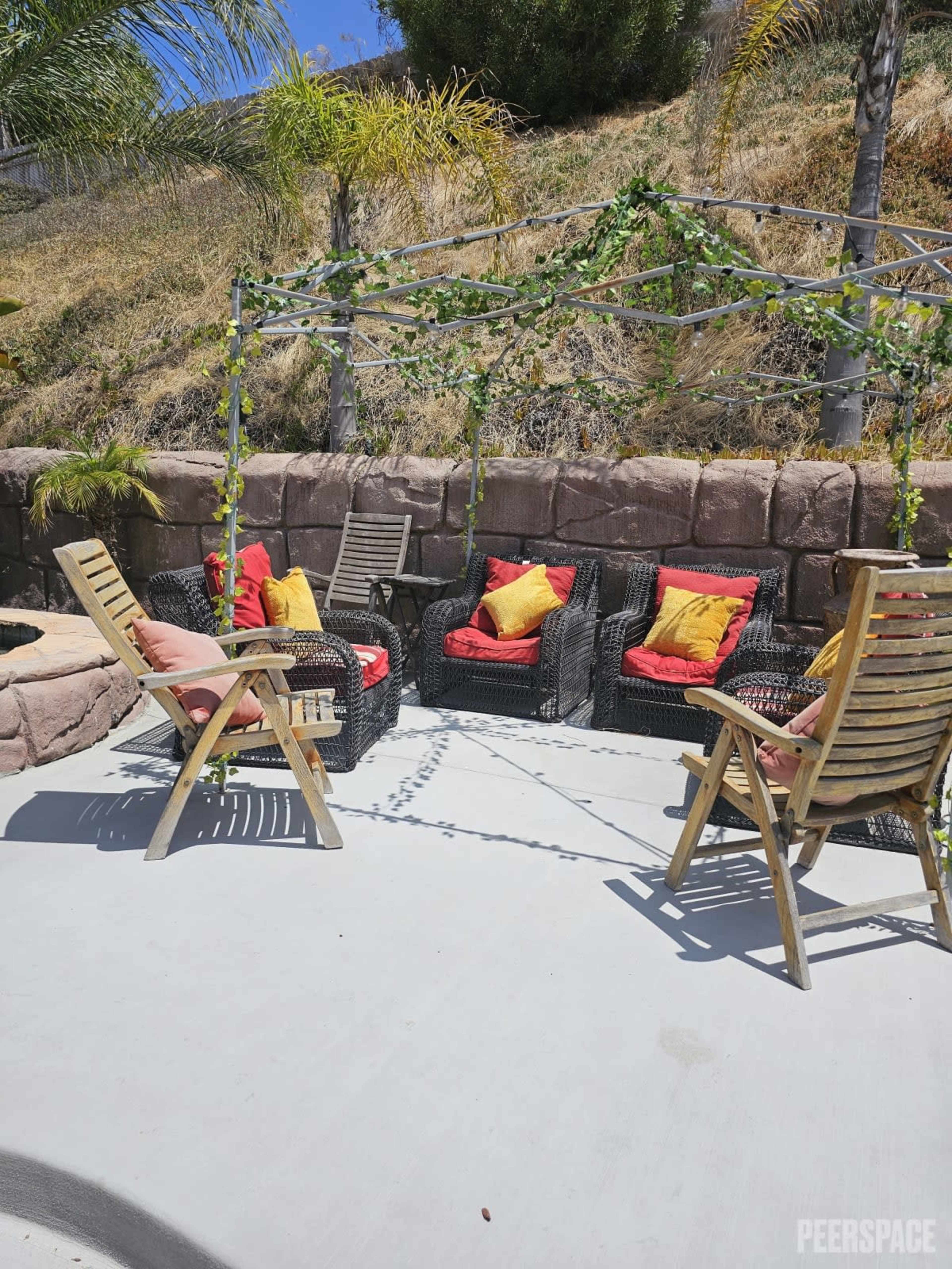 The scene features a modern outdoor seating area with two wooden chairs and a set of four cushioned chairs arranged under a vine-covered pergola.