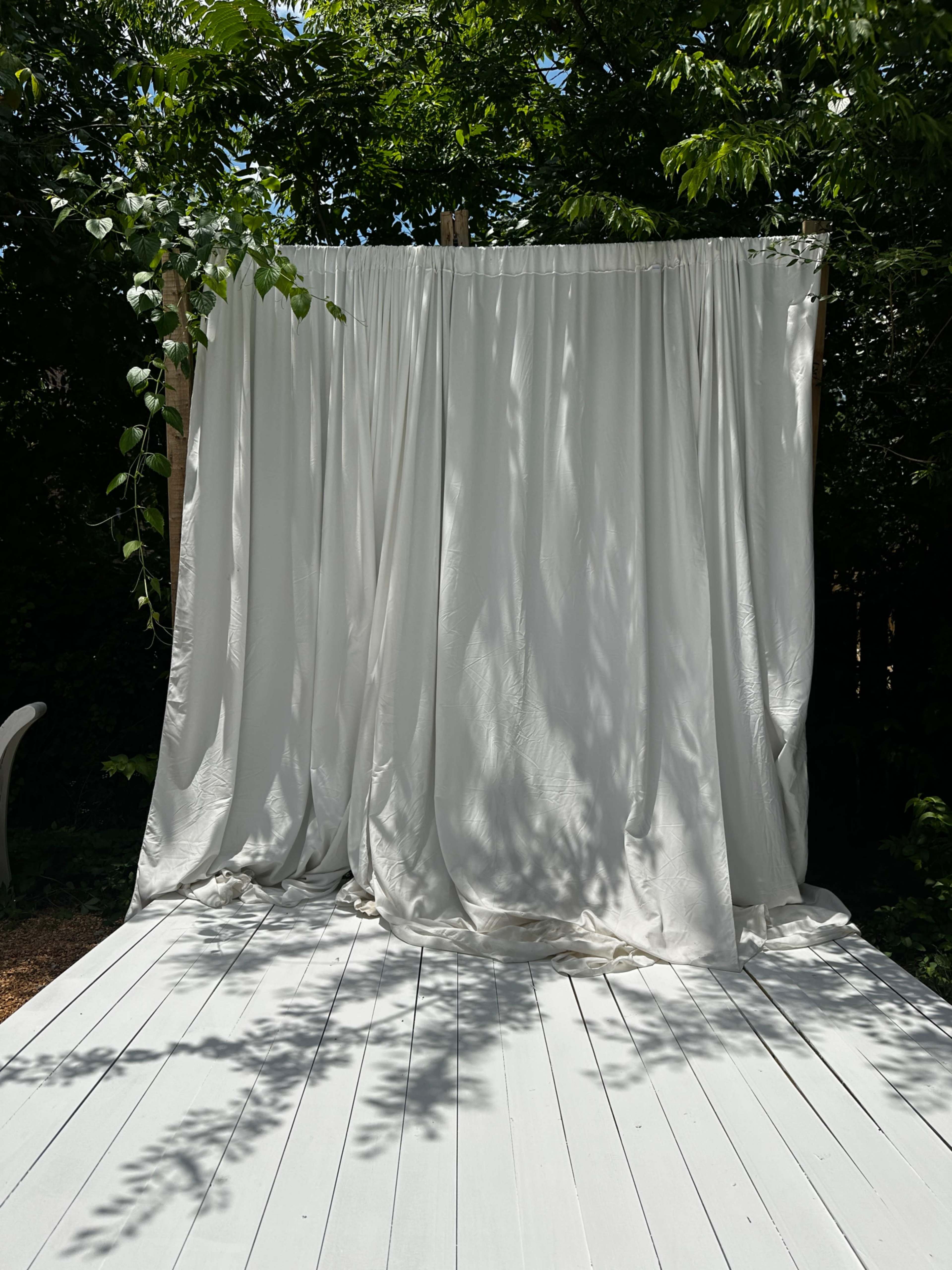 A white fabric curtain hangs on a wooden structure surrounded by greenery, with a white deck beneath it.