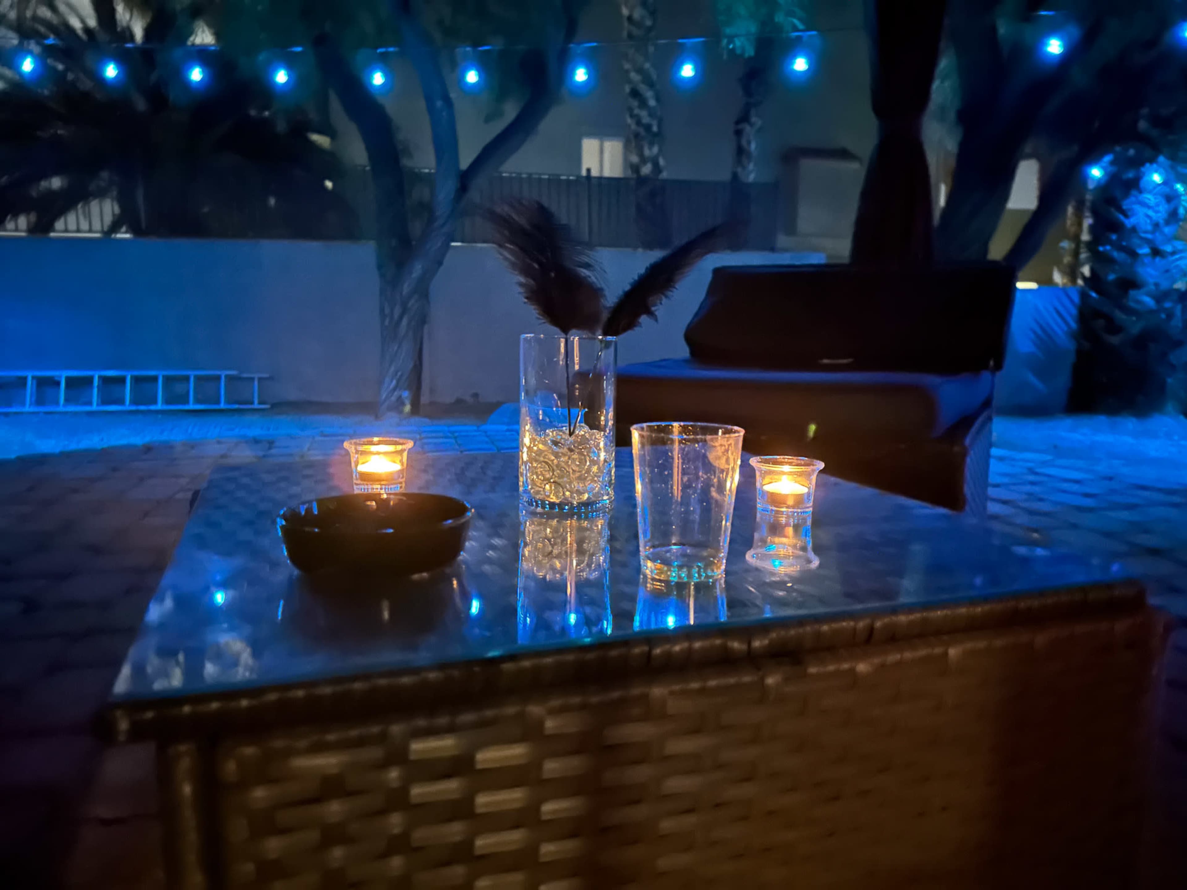 The image shows a table with candles and glassware, illuminated by blue lights in an outdoor setting surrounded by palm trees.