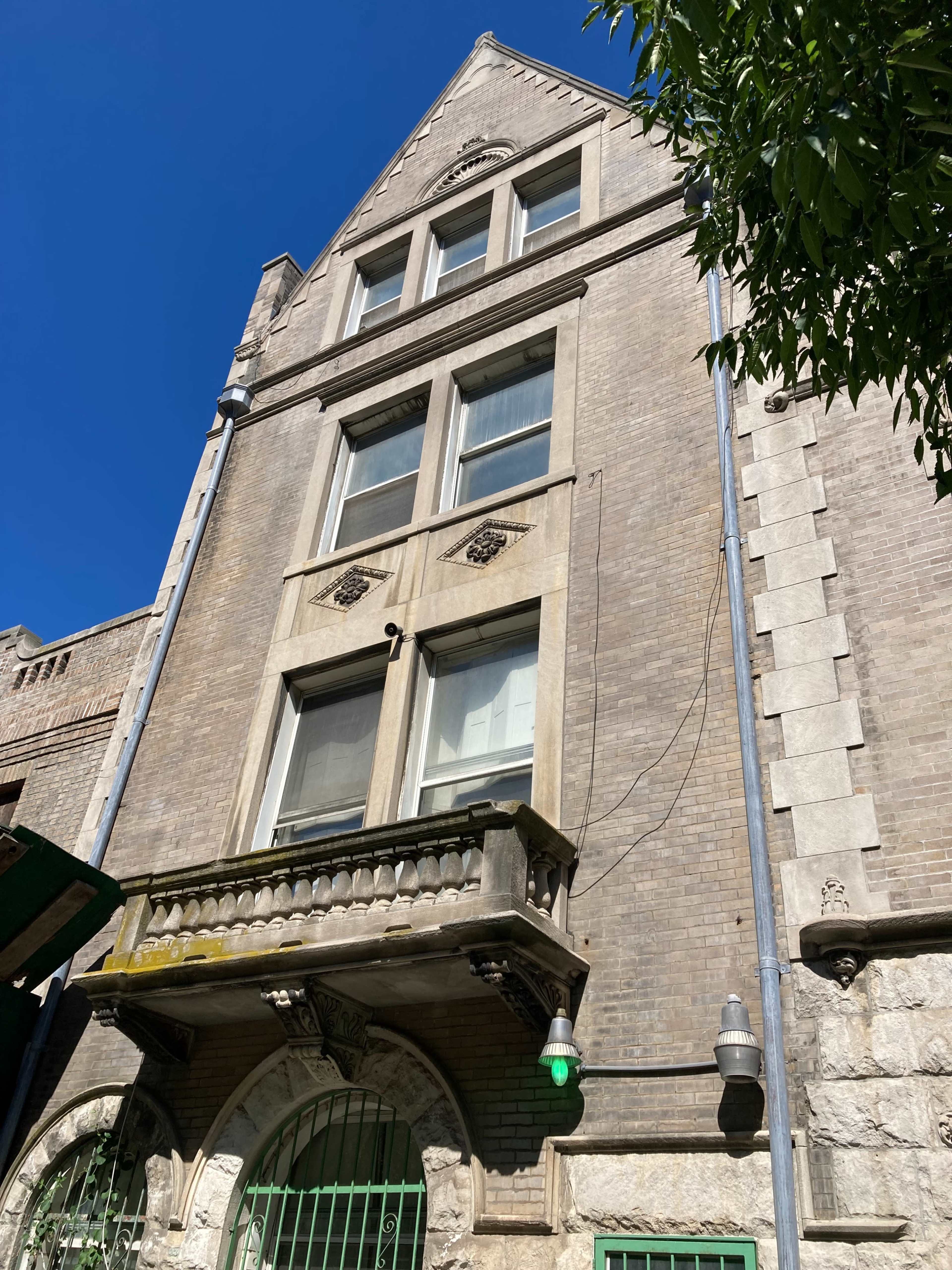 A tall, historic brick building with a peaked roof, multiple windows, and a balcony.