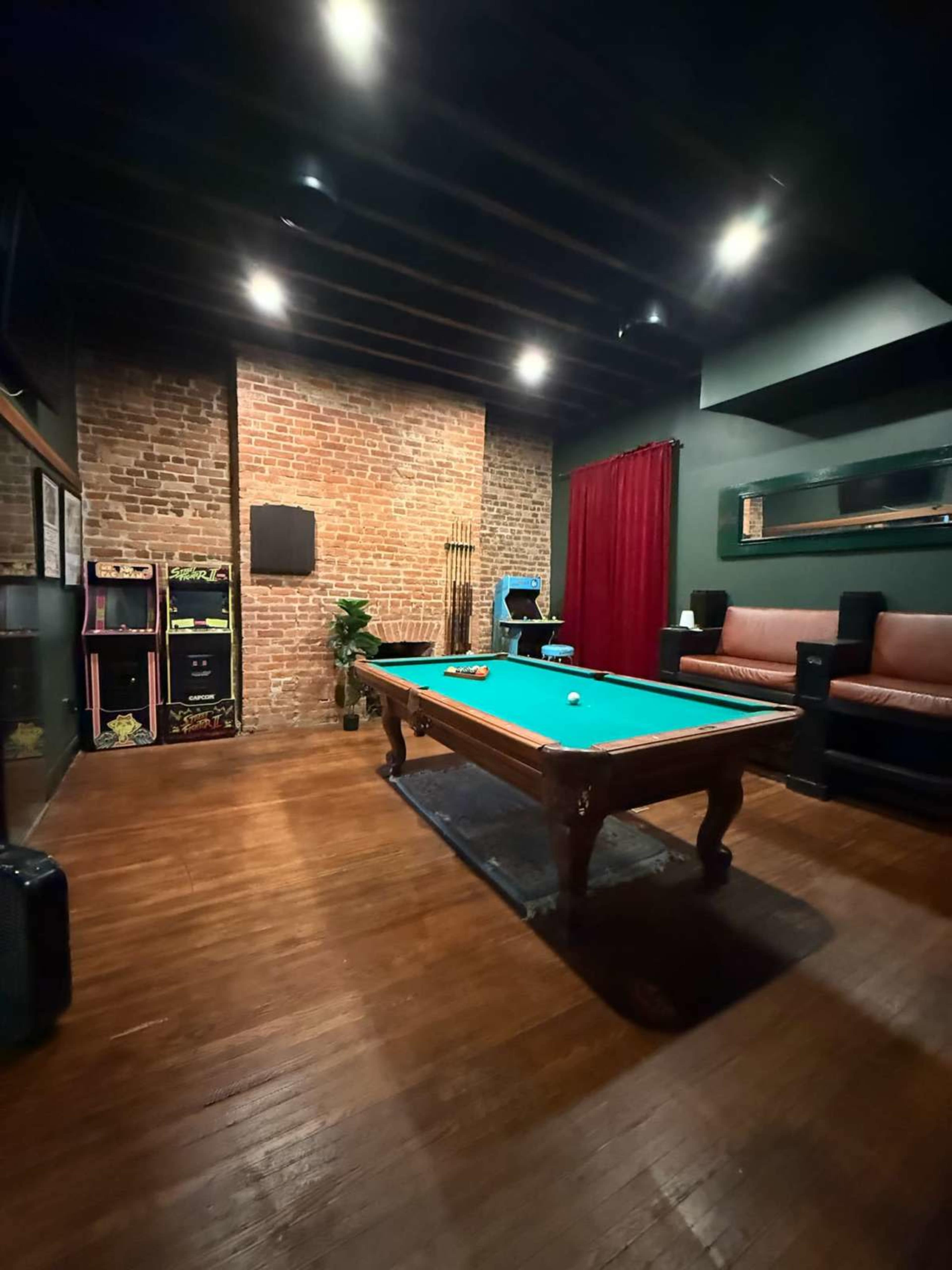 The image shows a game room with a green pool table, exposed brick walls, a dark wooden floor, and a seating area with a brown couch.