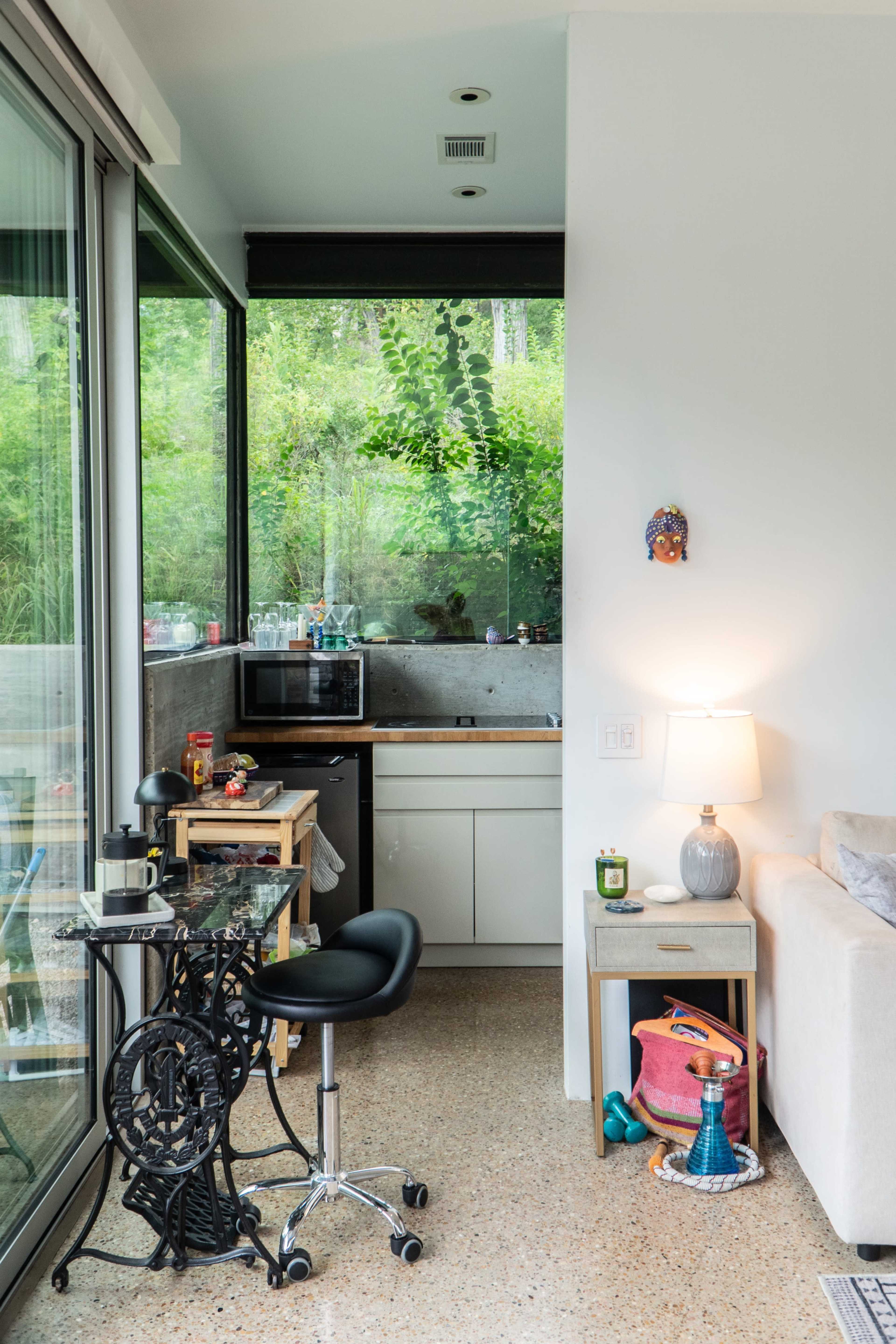 The image shows a small kitchen area with a microwave, a sink, and a work table beside a sofa, all set against a backdrop of greenery visible through large windows.