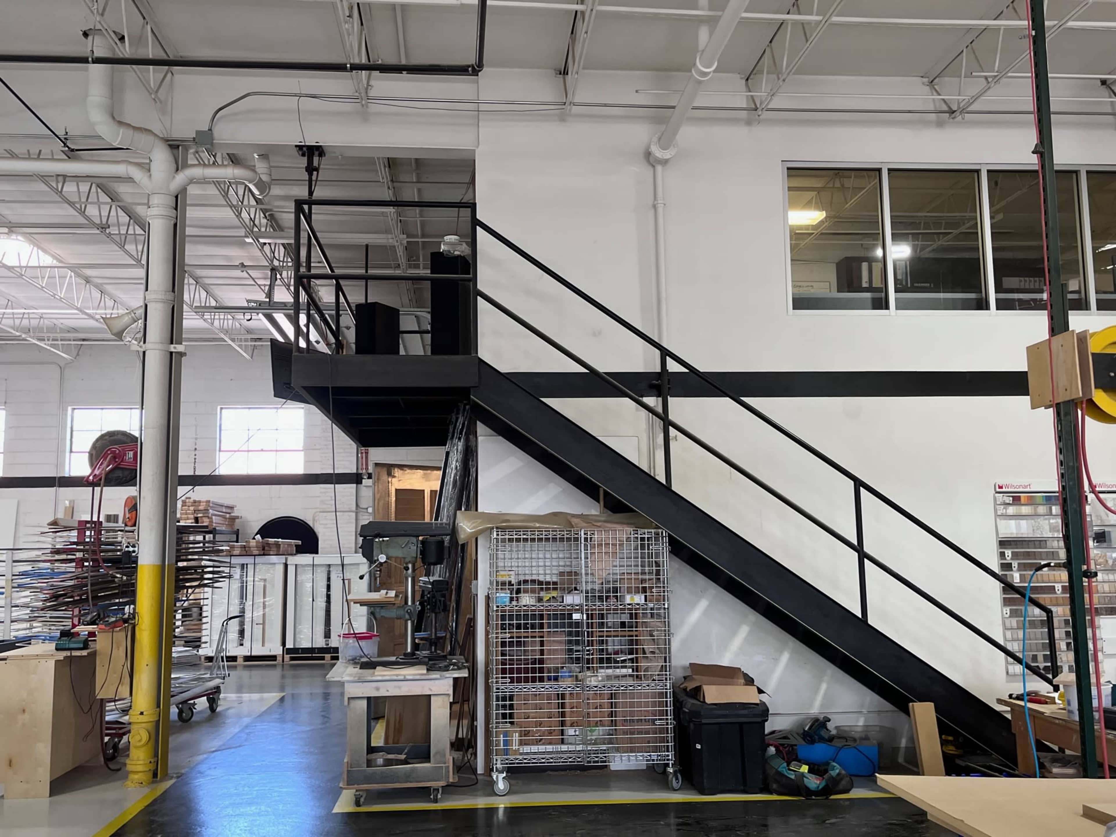 A warehouse interior with a black metal staircase leading to a second floor, alongside storage units and various materials on the ground level.