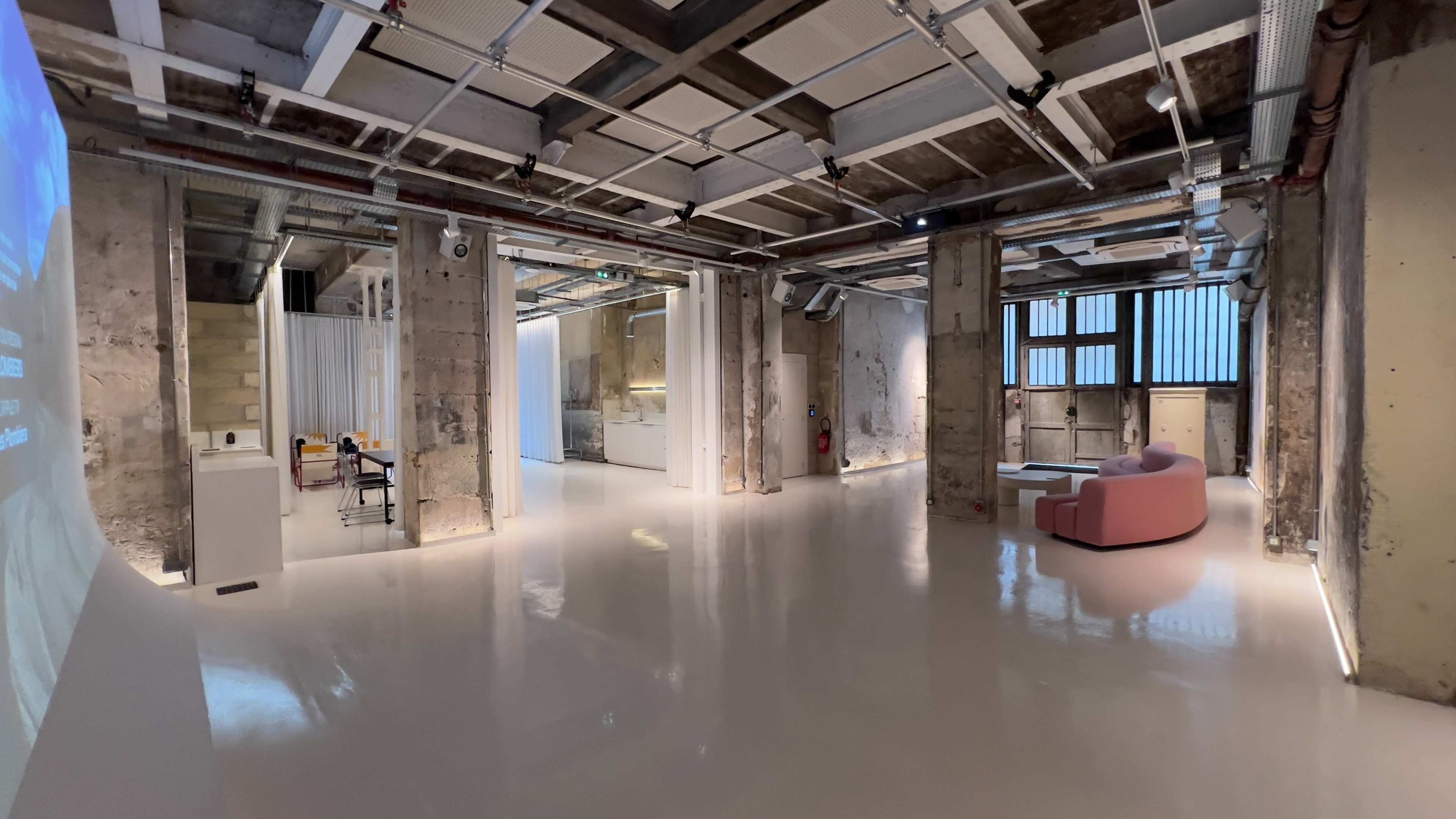 The image shows a spacious, modern interior with polished white floors, exposed concrete walls, and a seating area featuring a pink couch.
