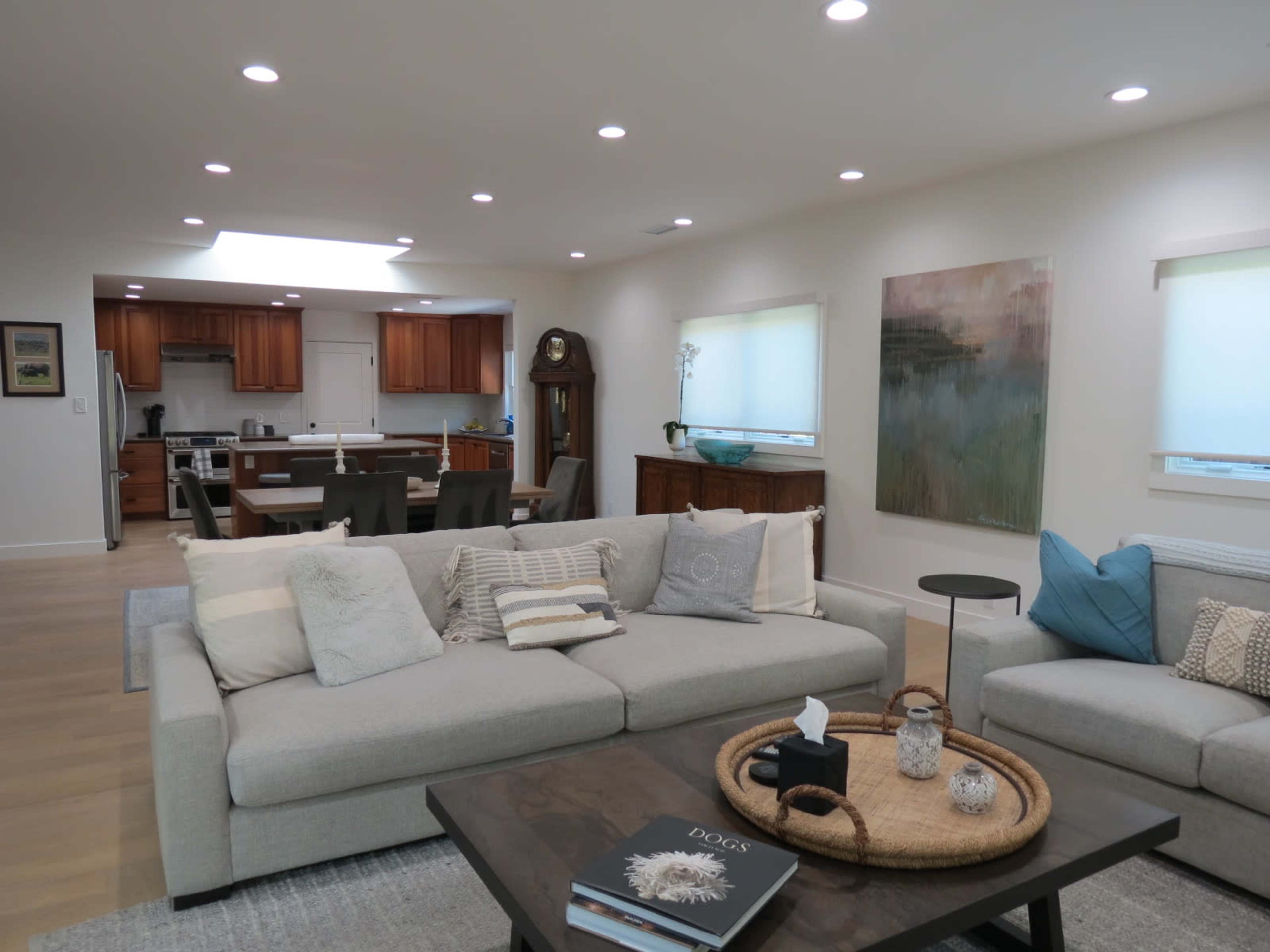 A modern living room features a light gray sectional sofa, a coffee table with decorative items, and an open layout leading to a kitchen area with wooden cabinets.