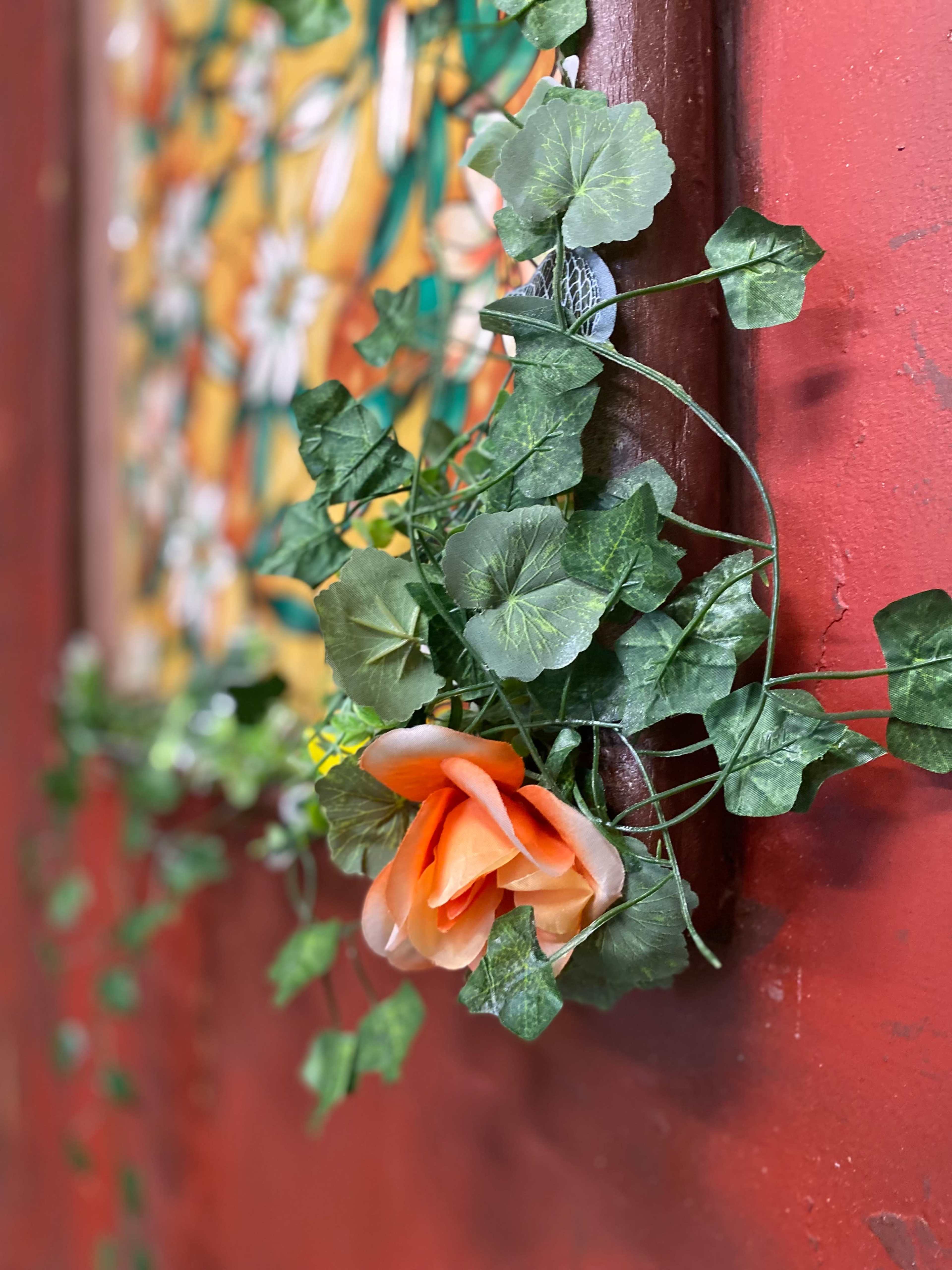 A plastic flower and green ivy leaves are arranged against a red wall.