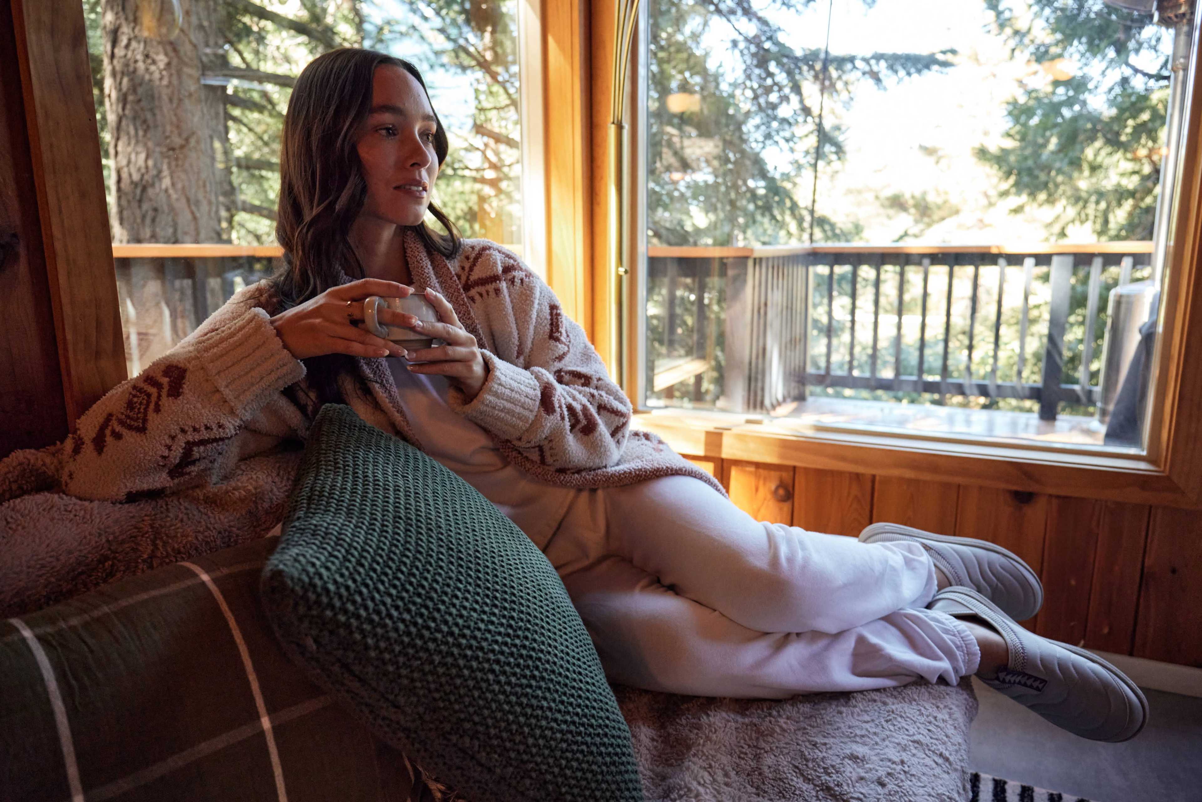 A woman sits comfortably on a couch in a cabin, holding a glass while looking out a large window surrounded by trees.