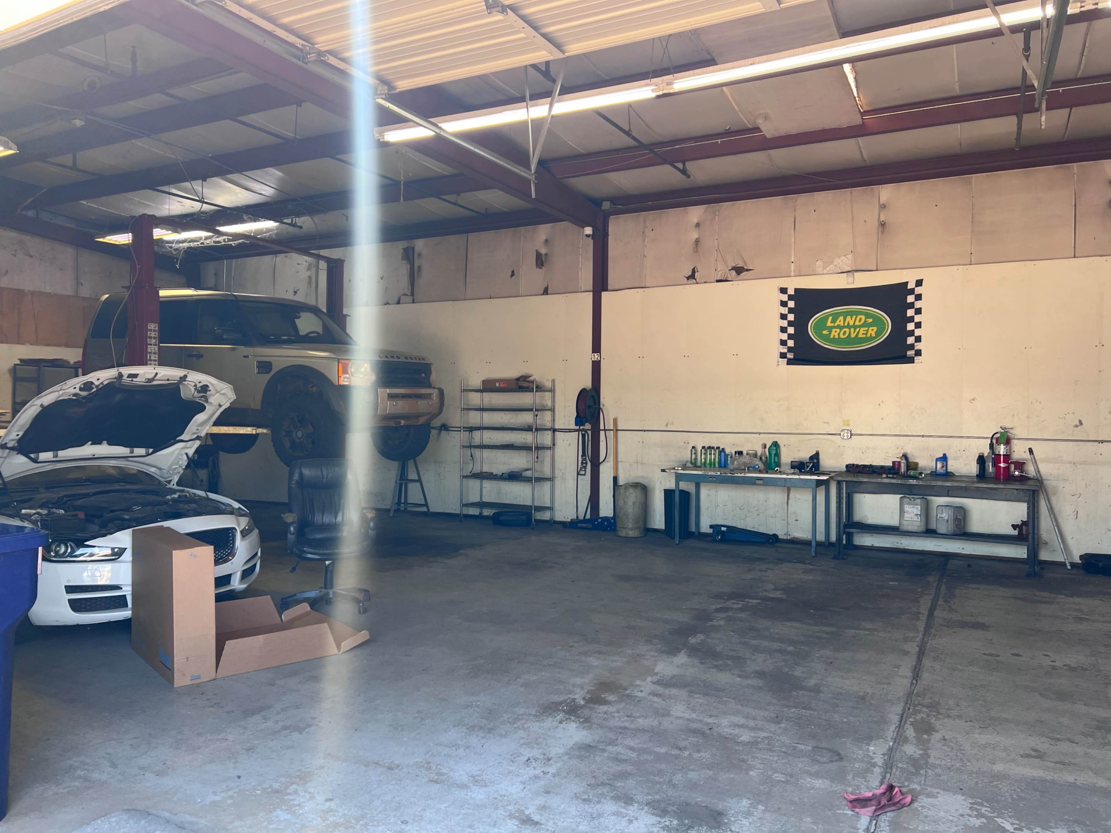 The image shows an automotive workshop with two vehicles, one on a lift and one parked with its hood open, alongside various tools and equipment arranged on shelves.