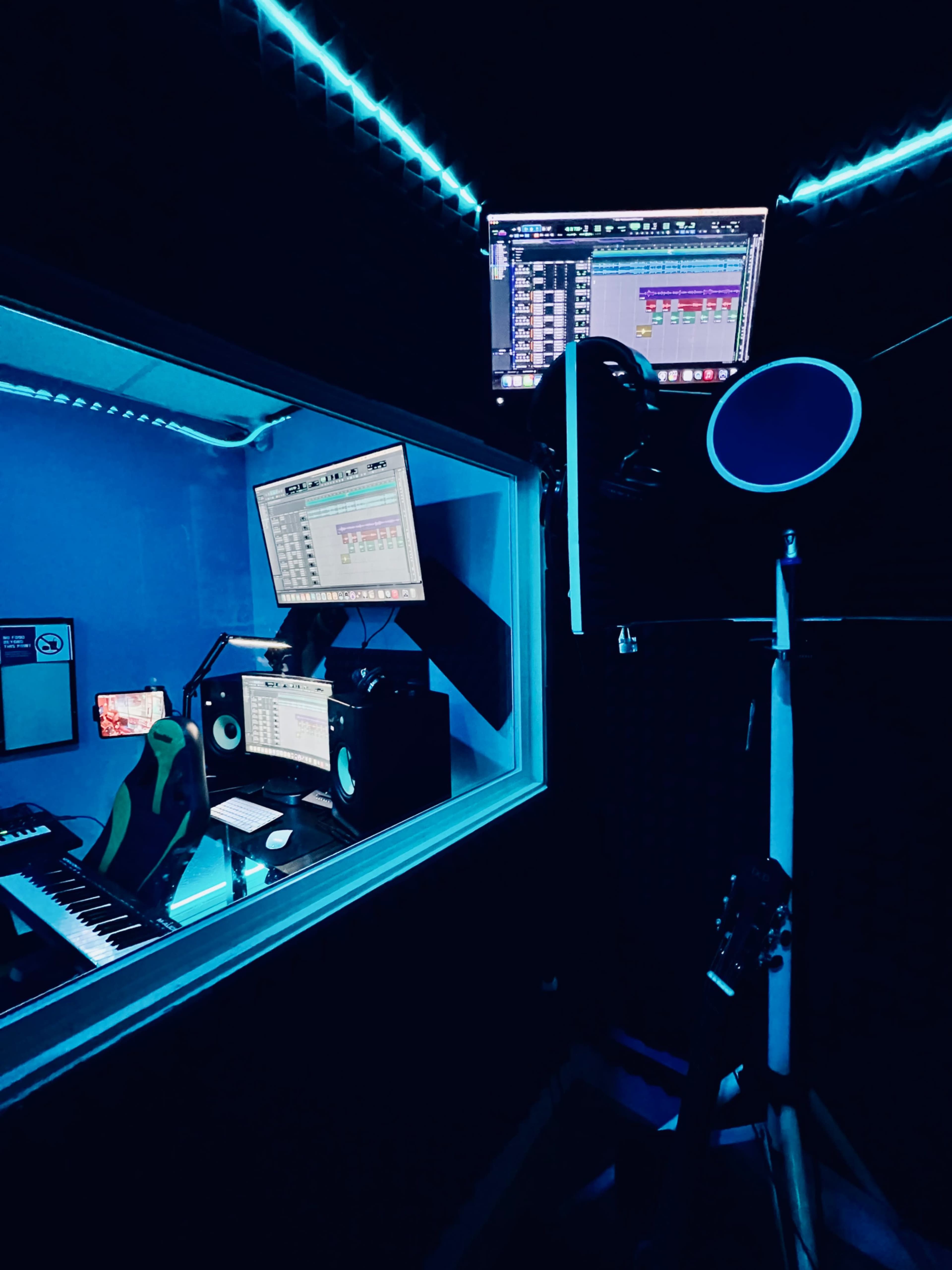A recording studio features a glass enclosure with two monitors displaying audio software, a keyboard, and various music equipment illuminated by blue lighting.