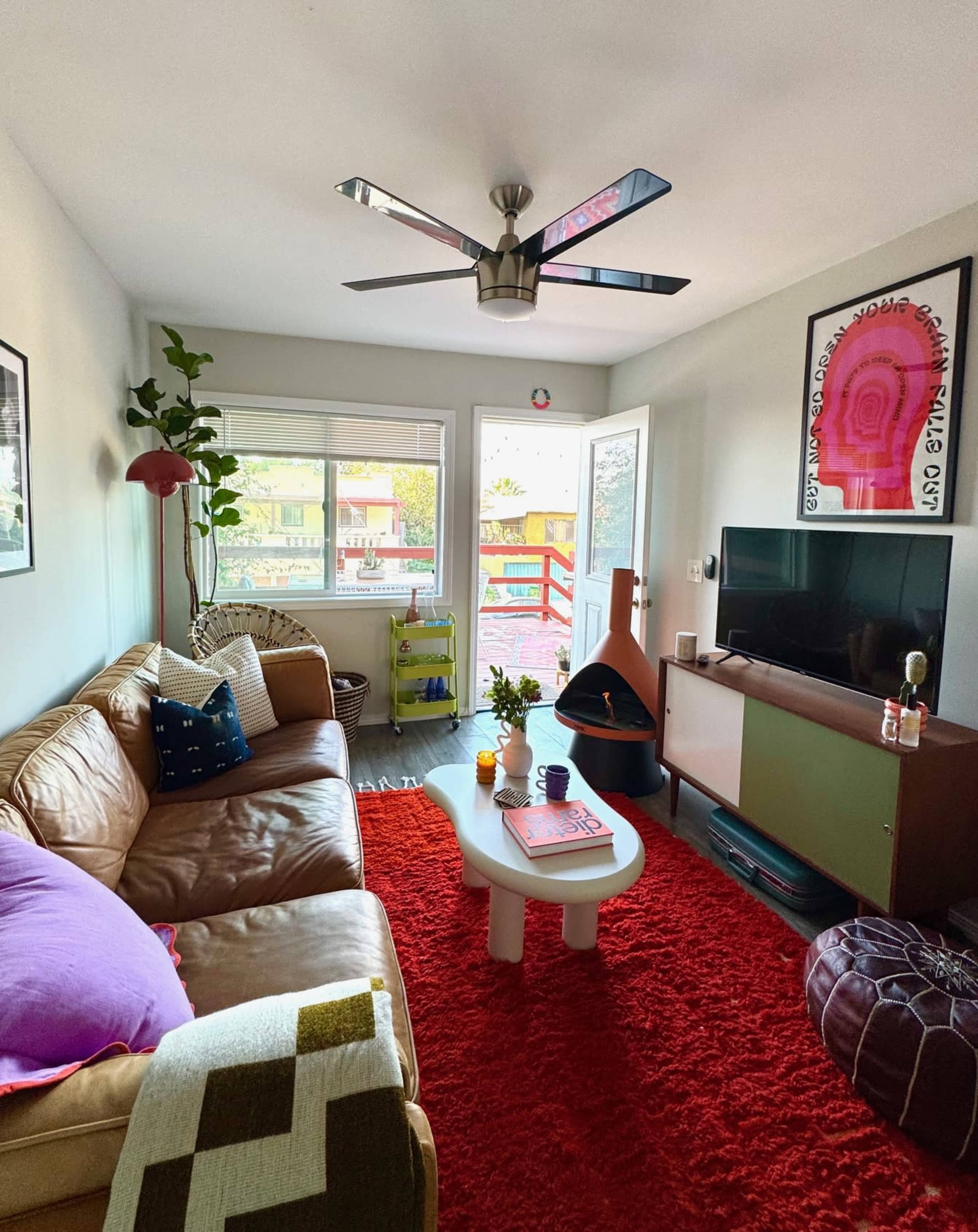 The image shows a cozy living room with a brown leather sofa, a red rug, a television, and a modern ceiling fan, all surrounded by large windows.