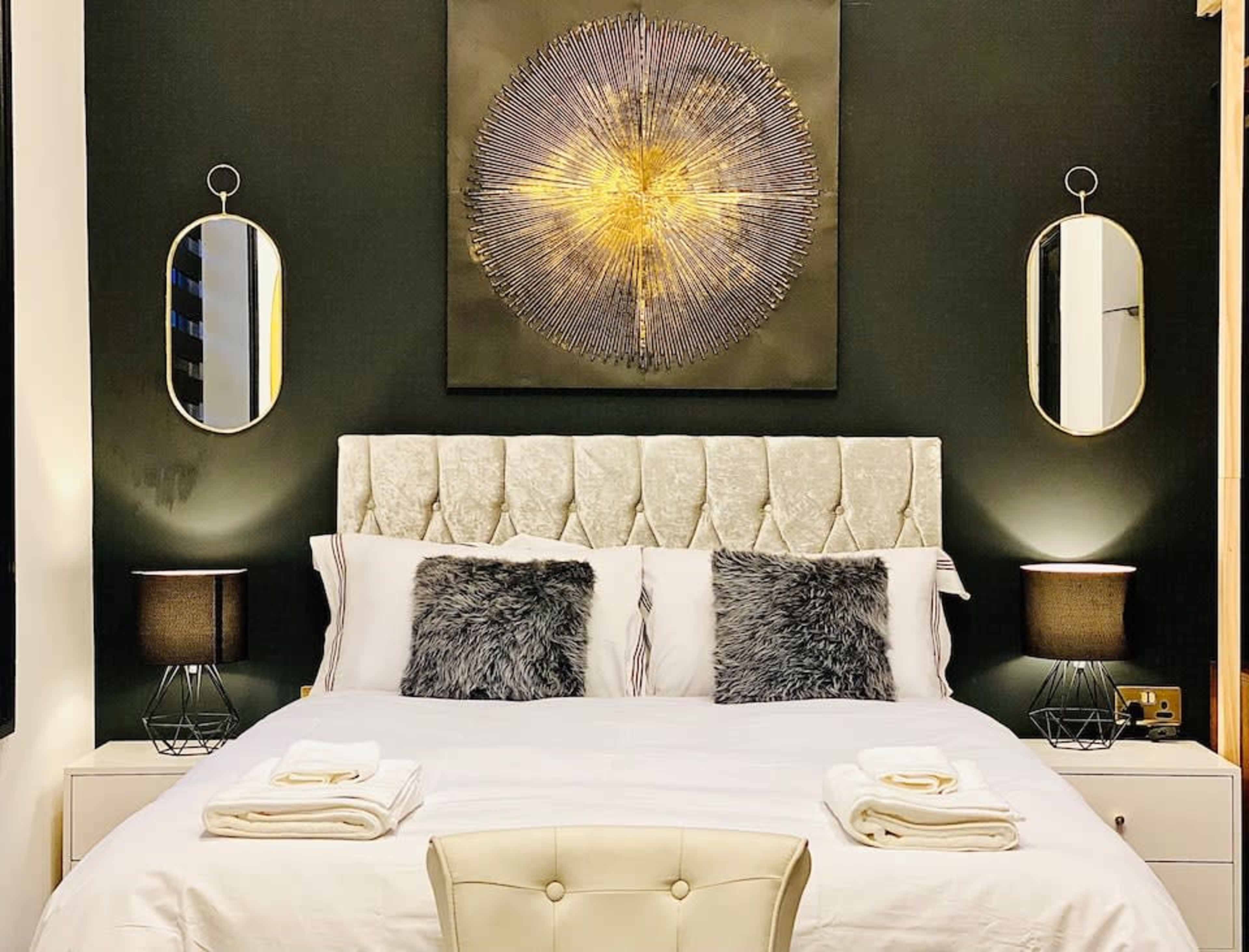 A neatly arranged bedroom with a tufted headboard, two bedside lamps, and decorative mirrors on dark walls.