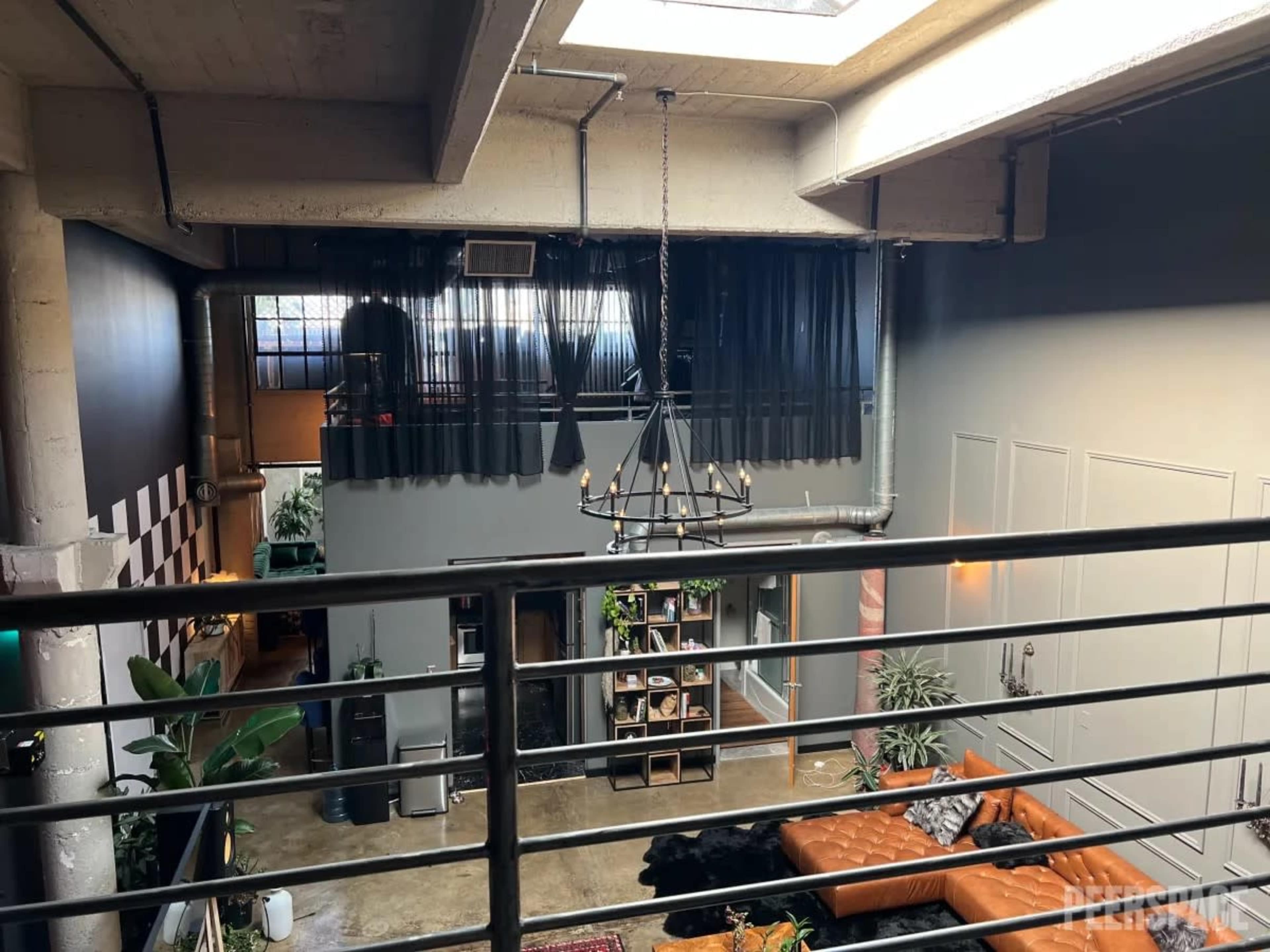 A multi-level loft space featuring a large chandelier, a modern orange couch, and a balcony with sheer black curtains.