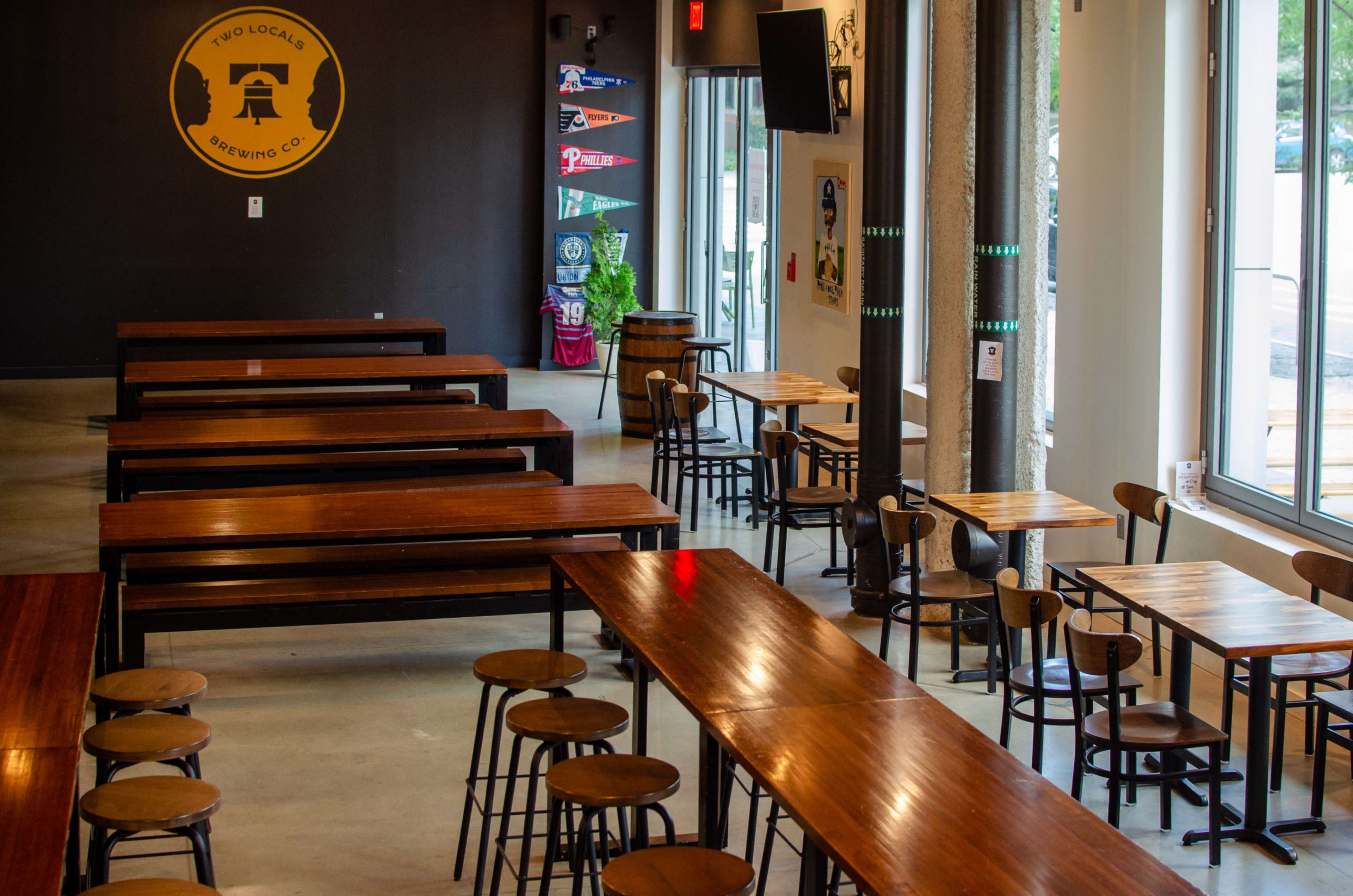 The image shows a spacious interior of a brewing company with long wooden tables and chairs, as well as wall decorations featuring various banners.