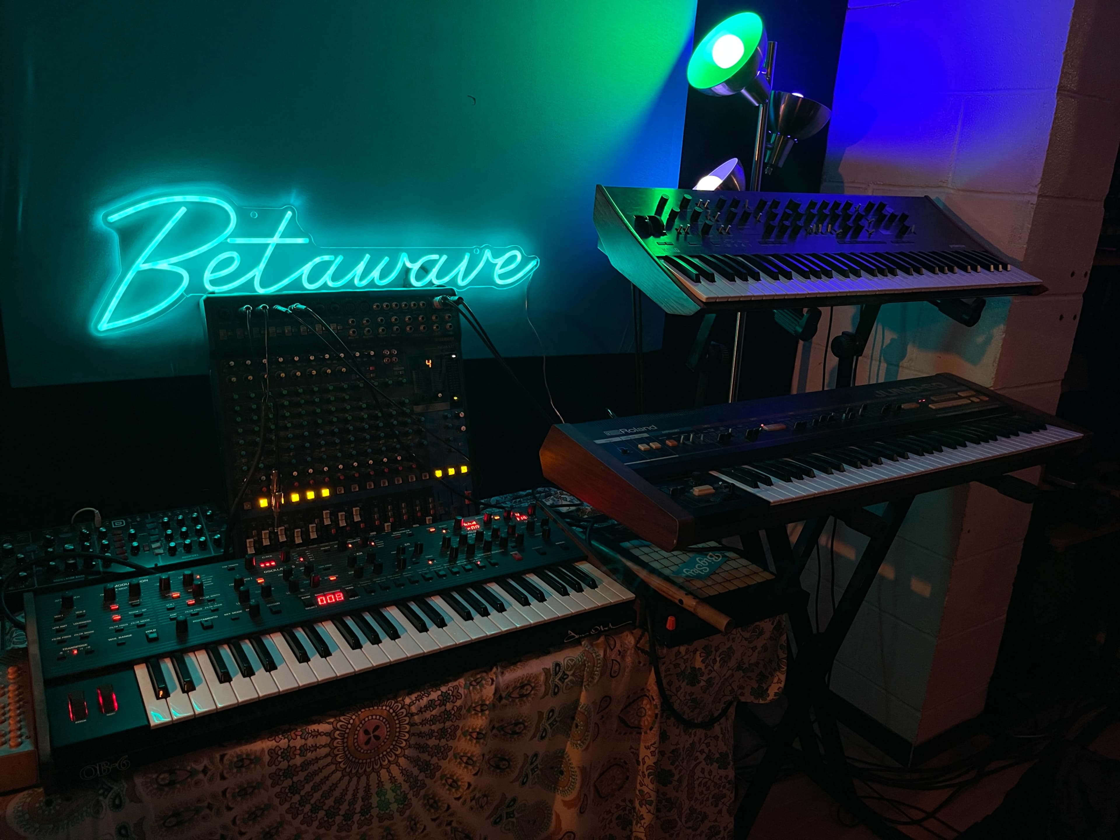 The image shows a collection of synthesizers and keyboards arranged in a music studio, illuminated by a neon "Betawave" sign.