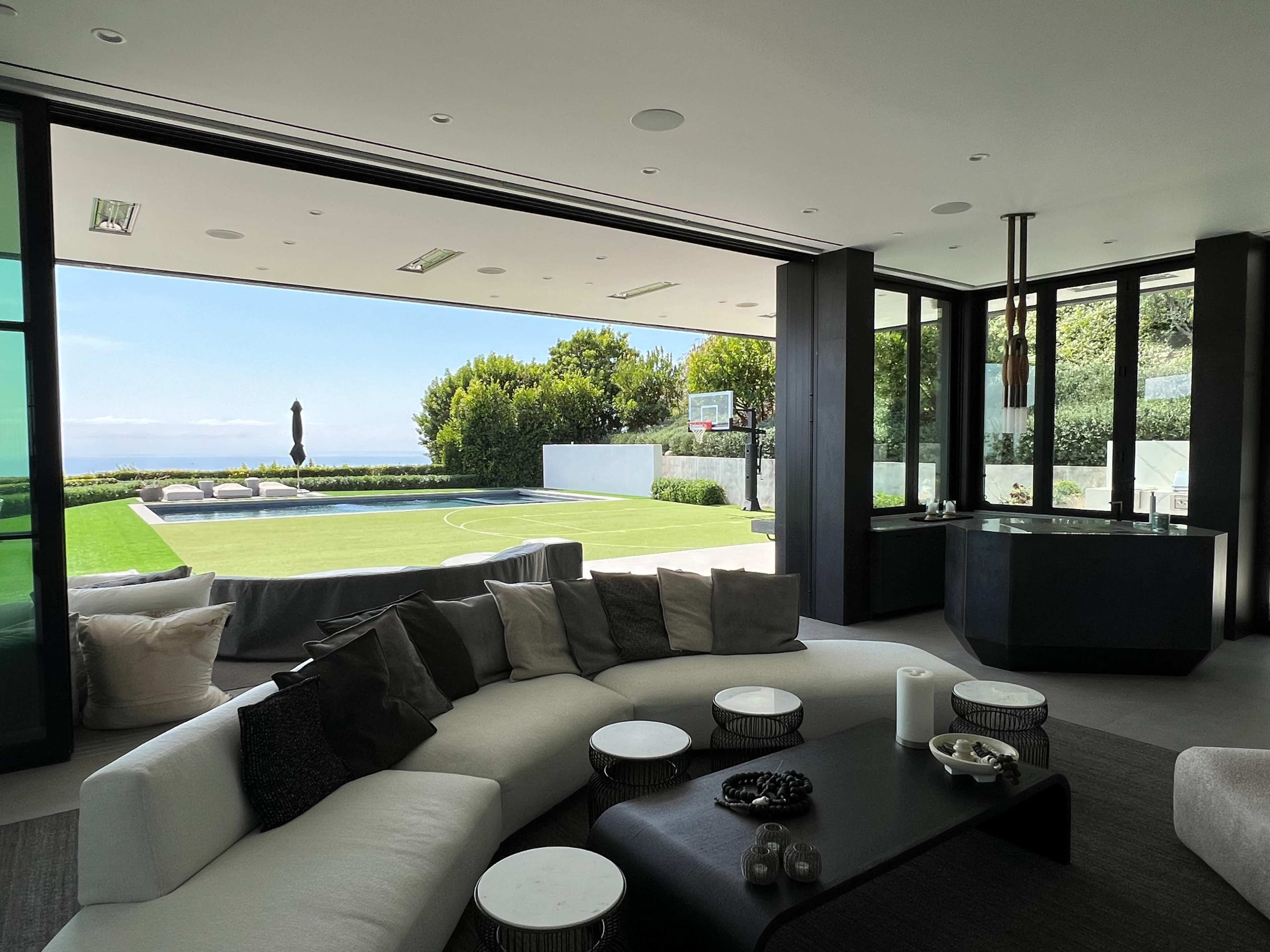 A modern living area features a circular white sofa, a dark coffee table, and large glass doors opening to a landscaped outdoor space with a basketball hoop and lounge seating.