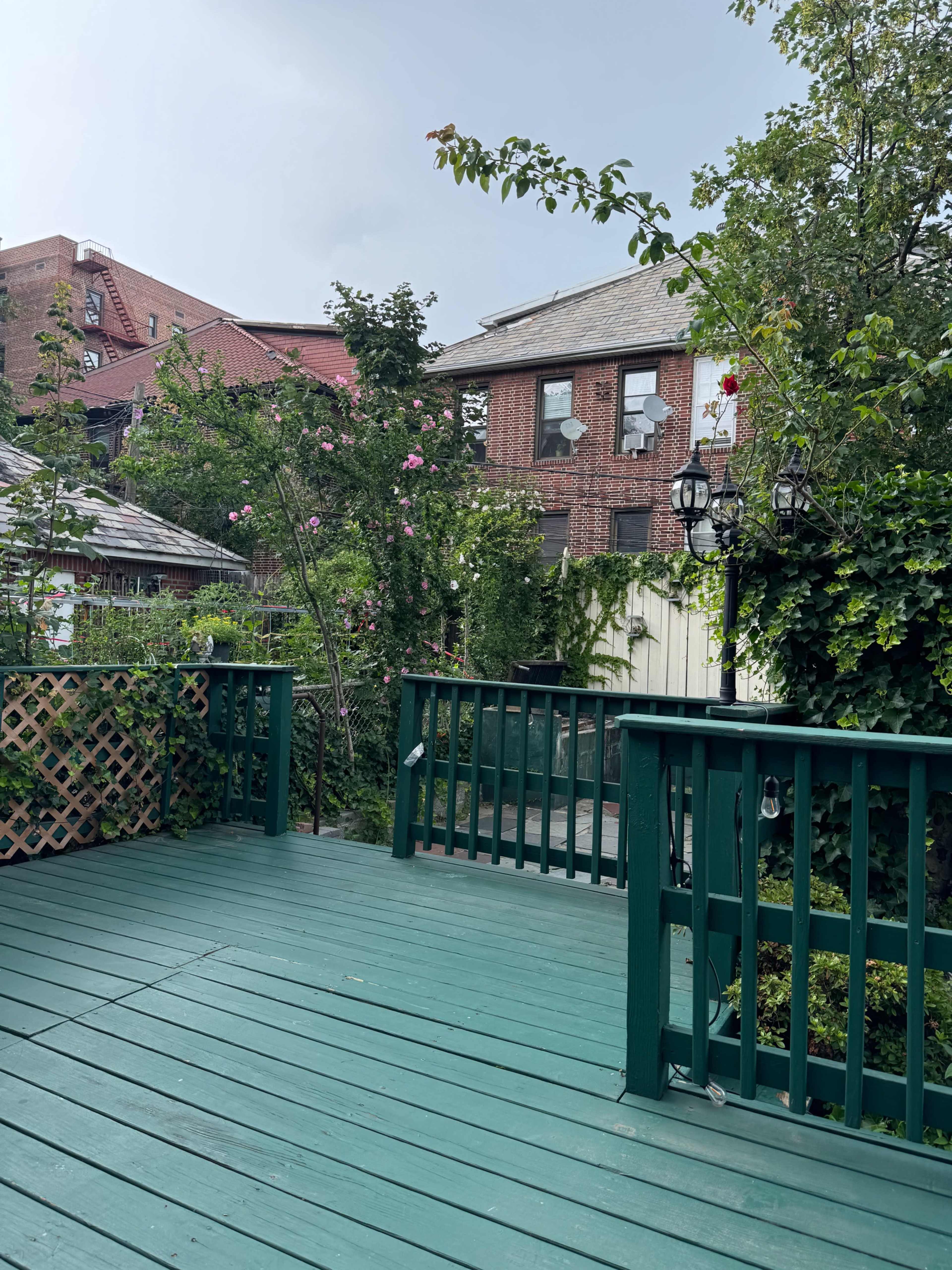 The image shows a green wooden deck surrounded by greenery and blooming flowers, with a brick building visible in the background.