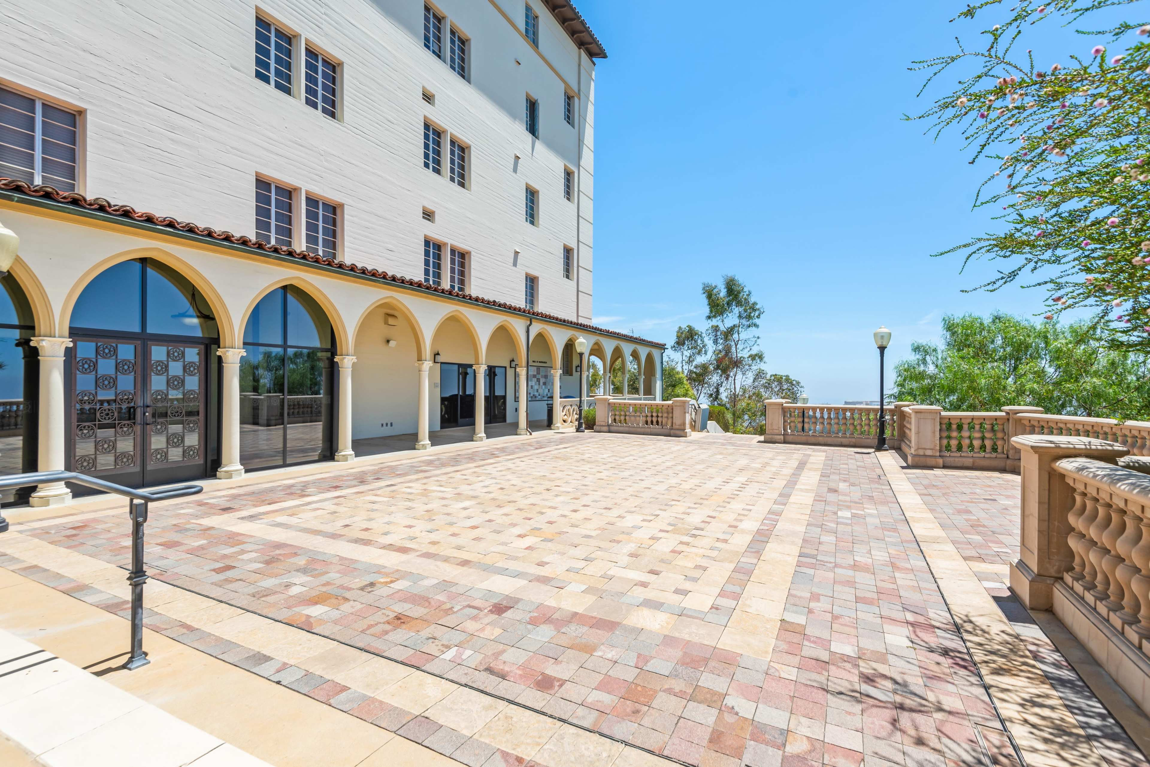 The image shows a spacious, paved courtyard in front of a multi-story building with arched doorways and large windows.