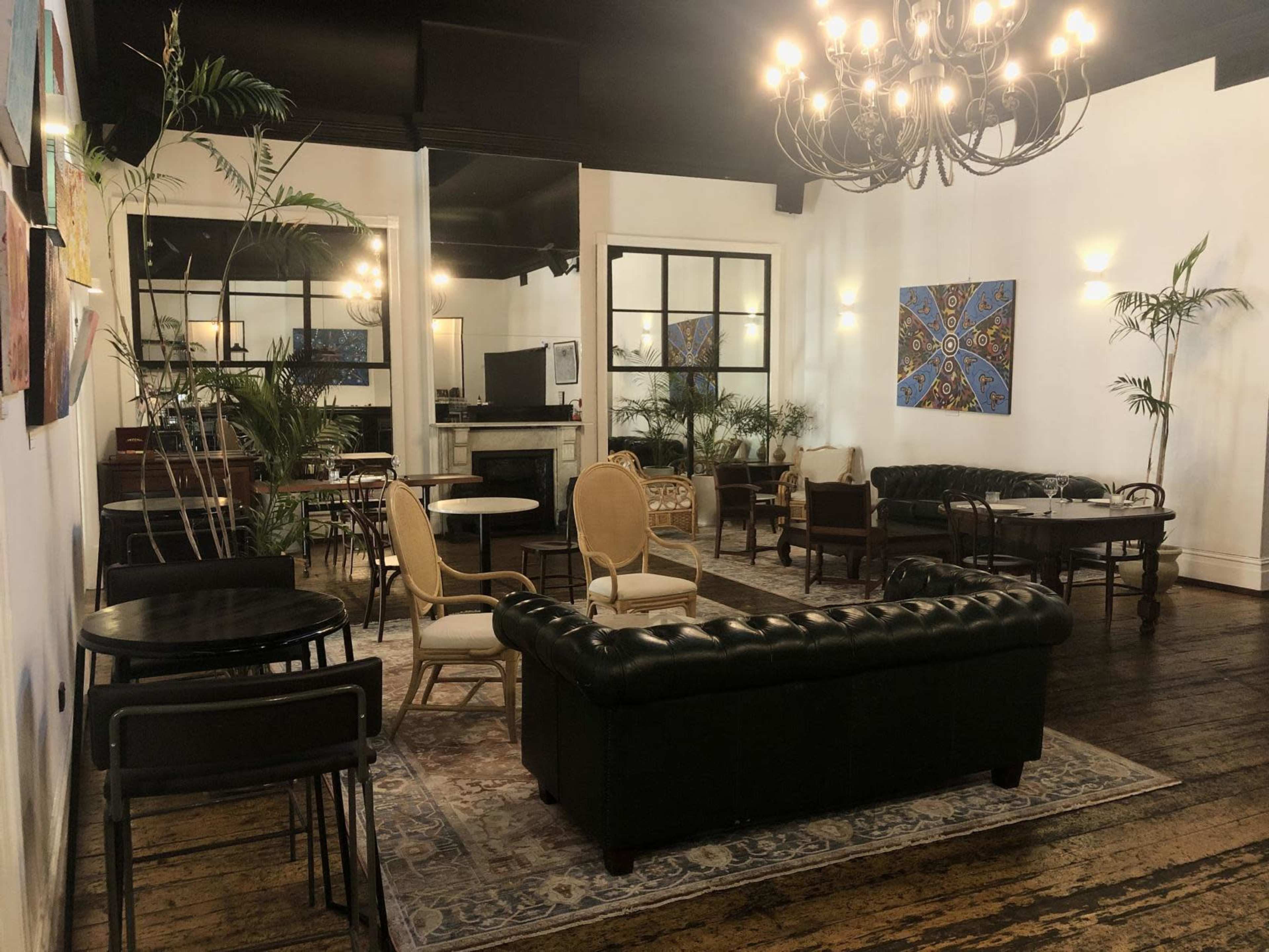 A stylish lounge area with various seating arrangements, including a leather sofa, wooden tables, and potted plants, set against a backdrop of white walls and artwork.