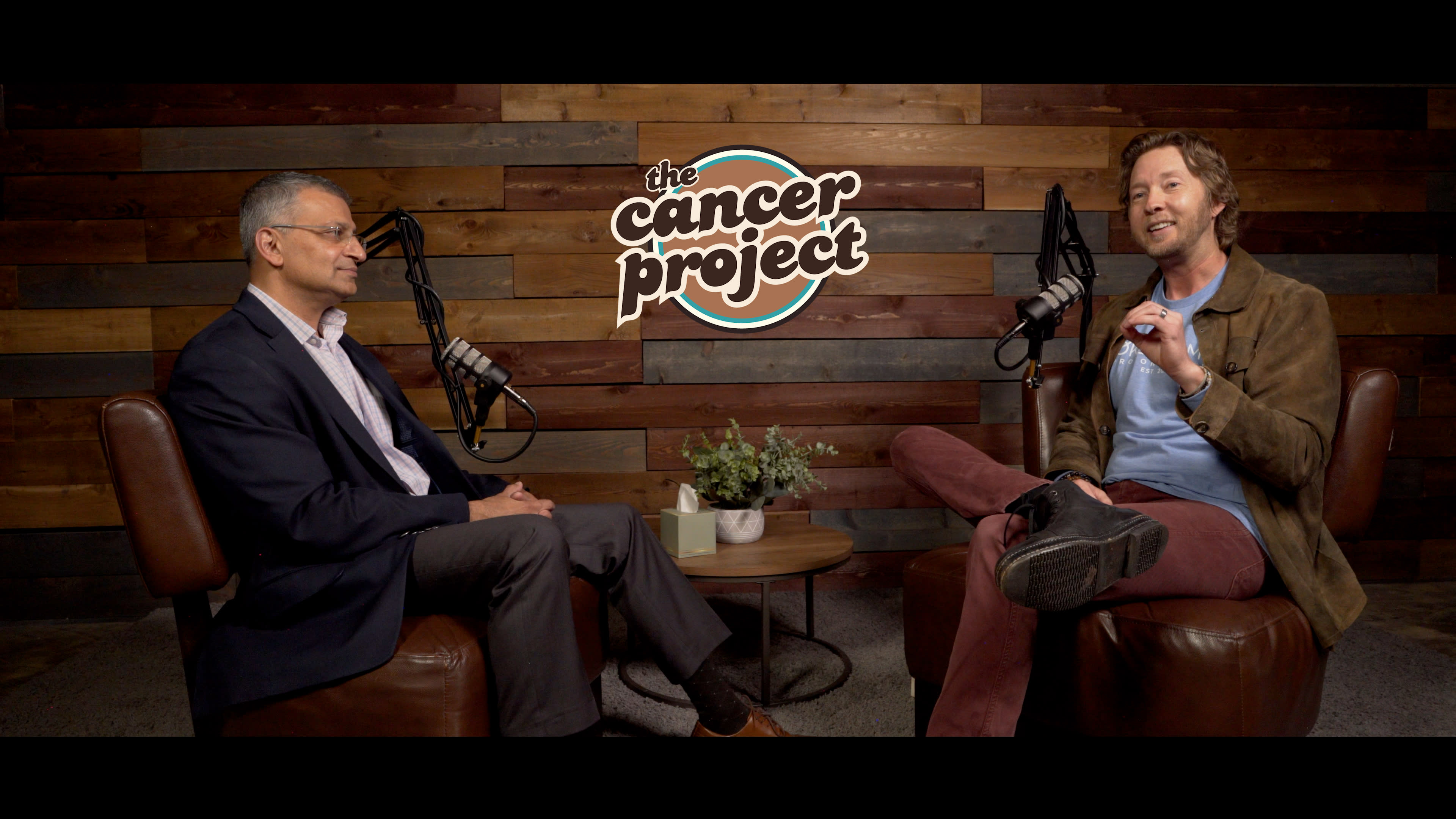 Two men are seated in armchairs in a studio setting, with microphones in front of them and a logo for "The Cancer Project" displayed in the background.
