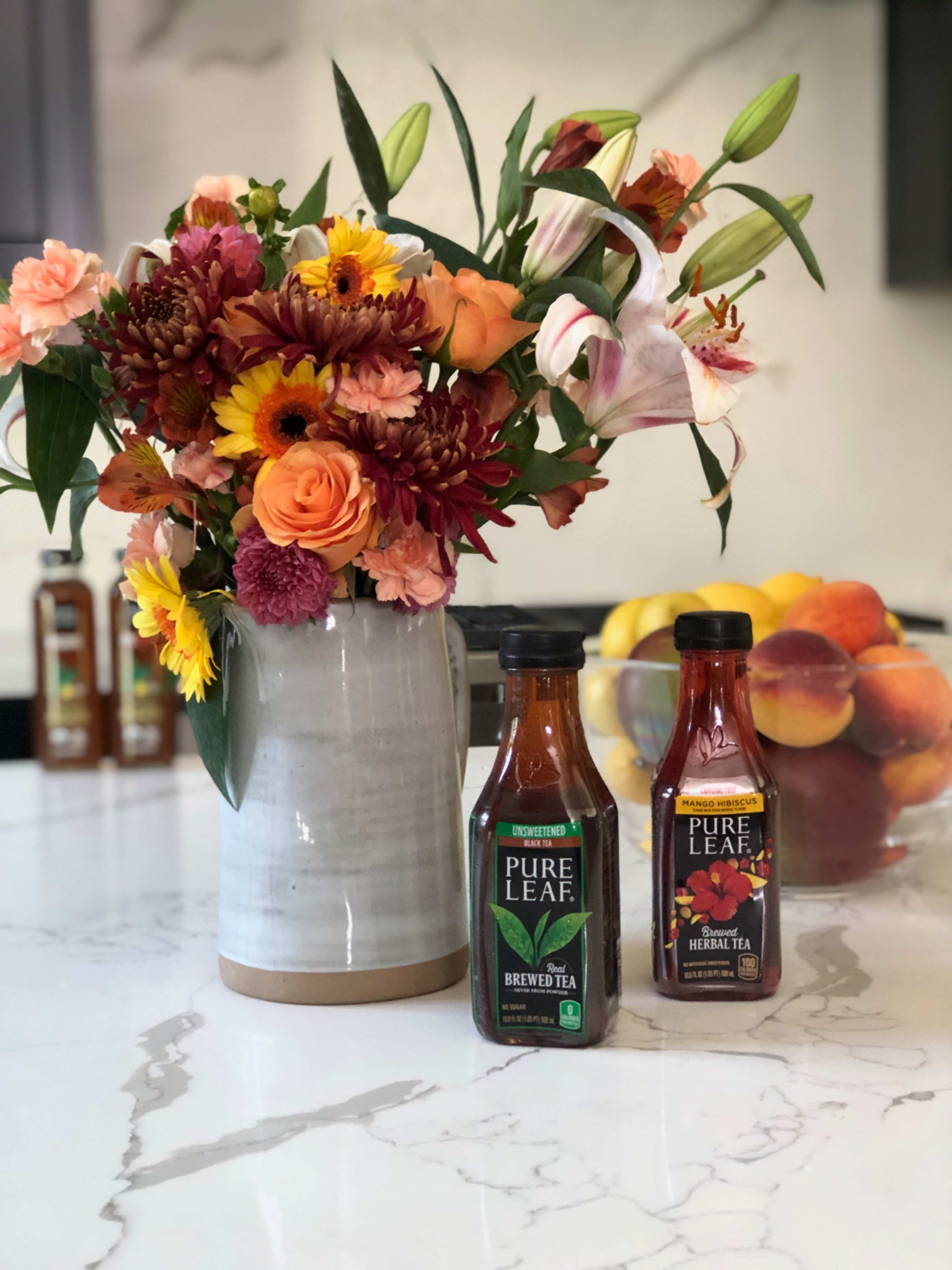 A vase filled with colorful flowers stands next to two bottles of Pure Leaf tea on a marble countertop, with a bowl of fruit in the background.