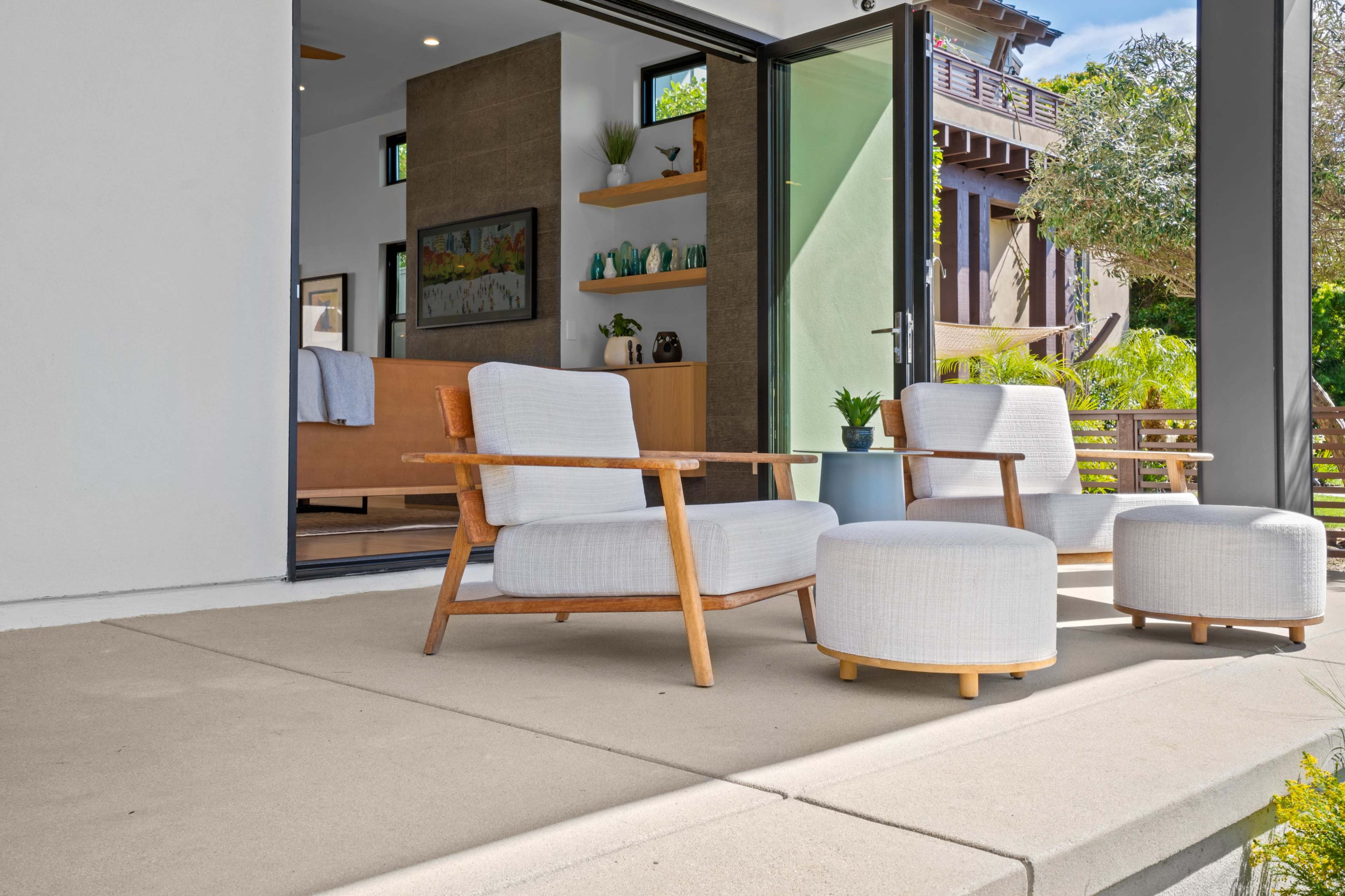 A modern outdoor seating area features two armchairs and a round ottoman placed on a concrete patio, with doors leading into a bright indoor space.