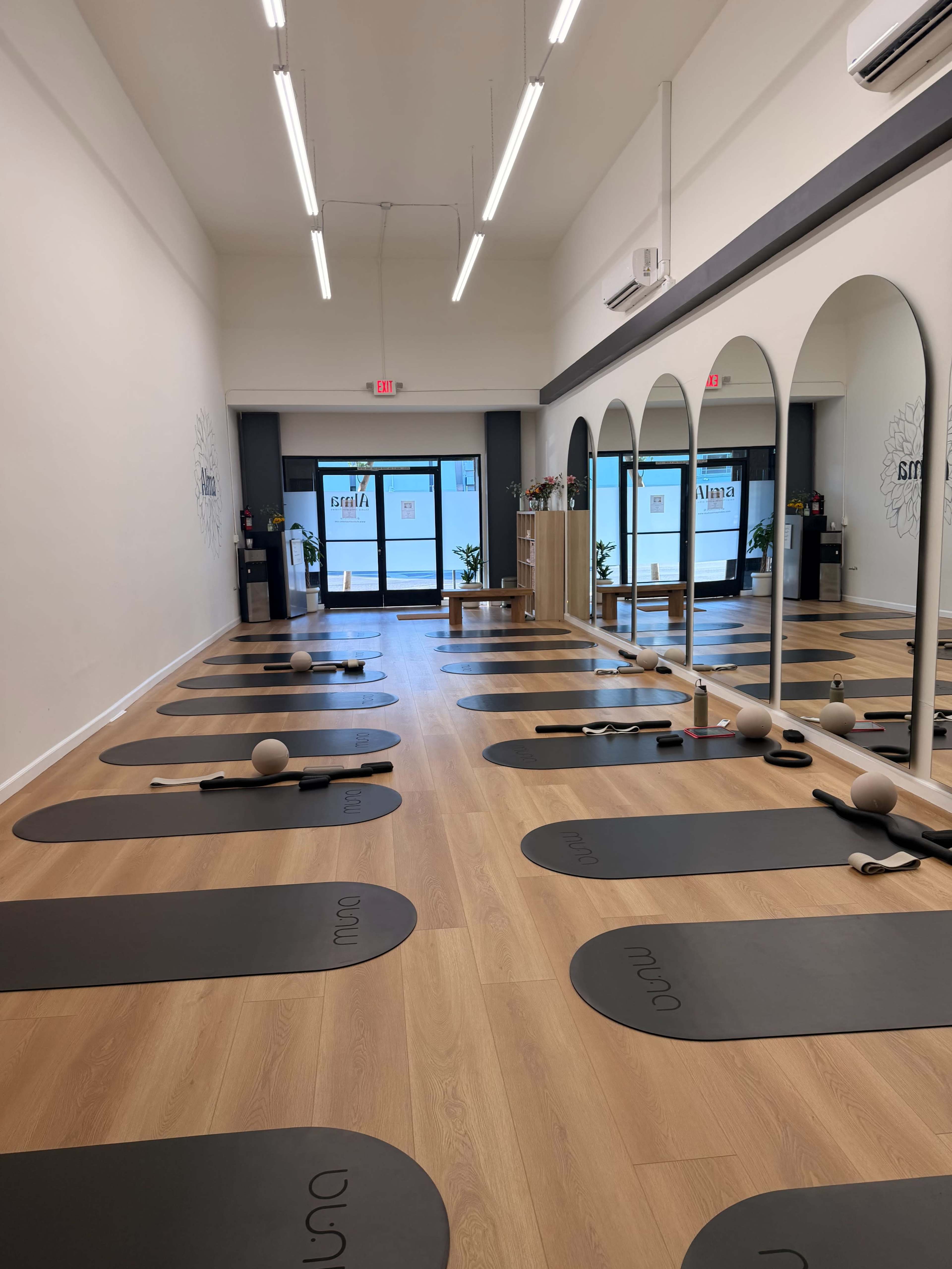A yoga studio features several mats lined up on a wooden floor, with a large mirror and arched doorways leading to the entrance.