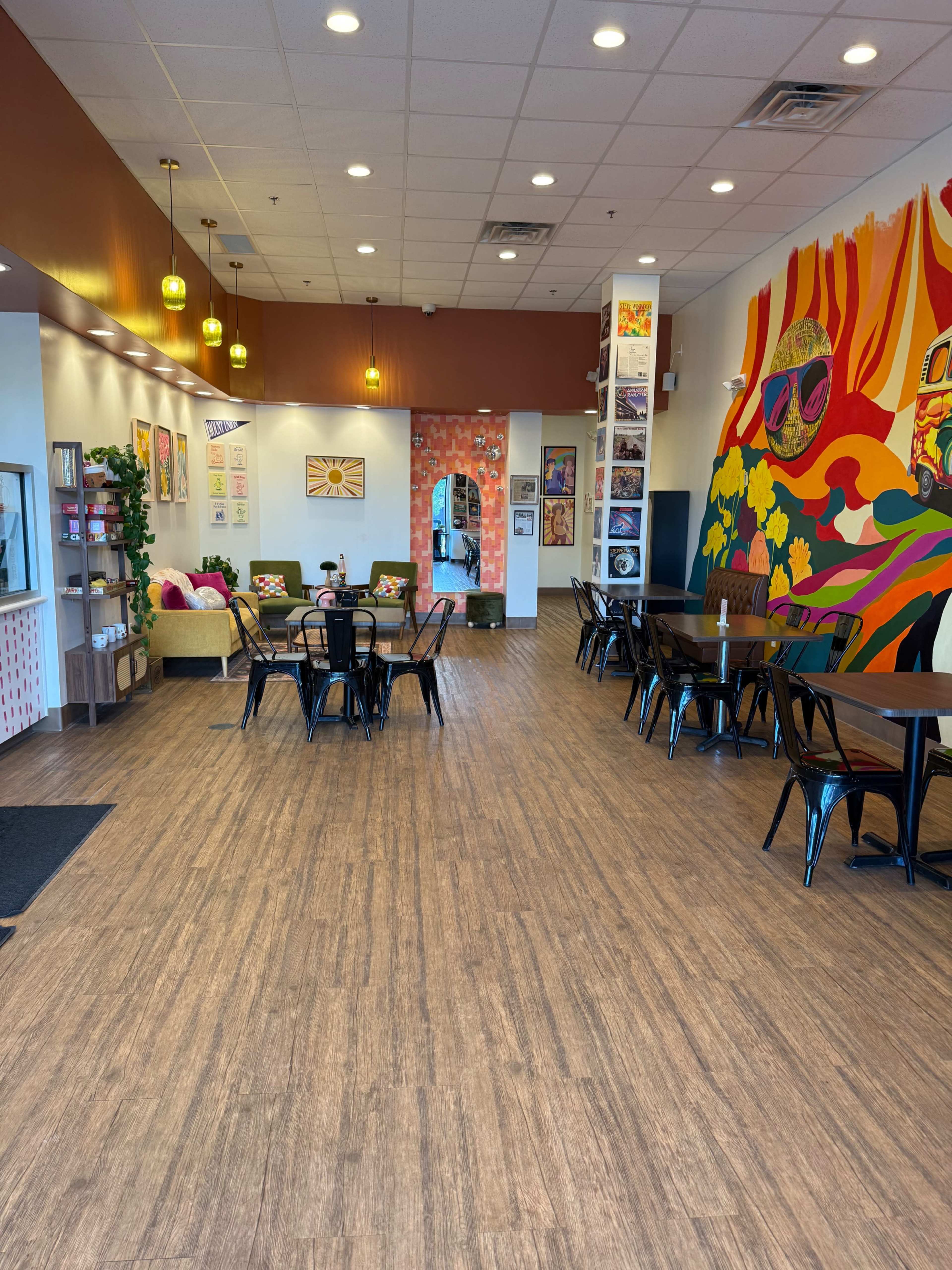 The image shows a brightly lit café with wooden flooring, colorful wall art, and a mixture of seating arrangements including tables and a lounge area.