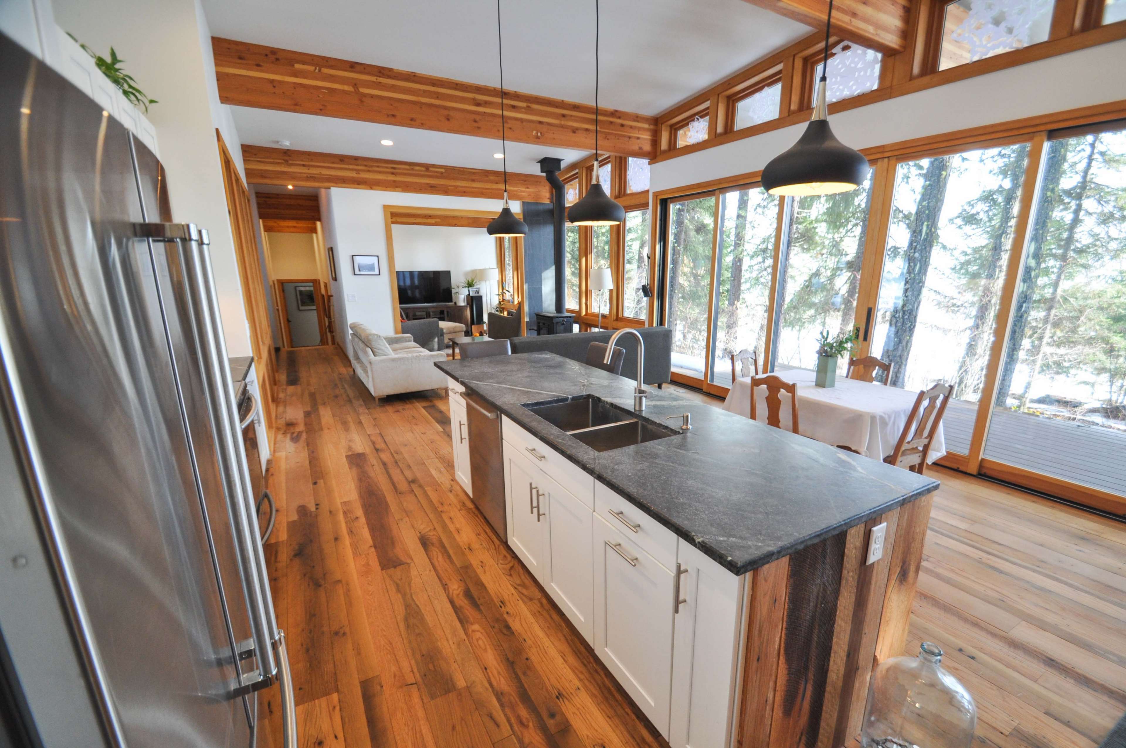 The image shows a modern kitchen with a central island, stainless steel appliances, and large windows overlooking a wooded area.