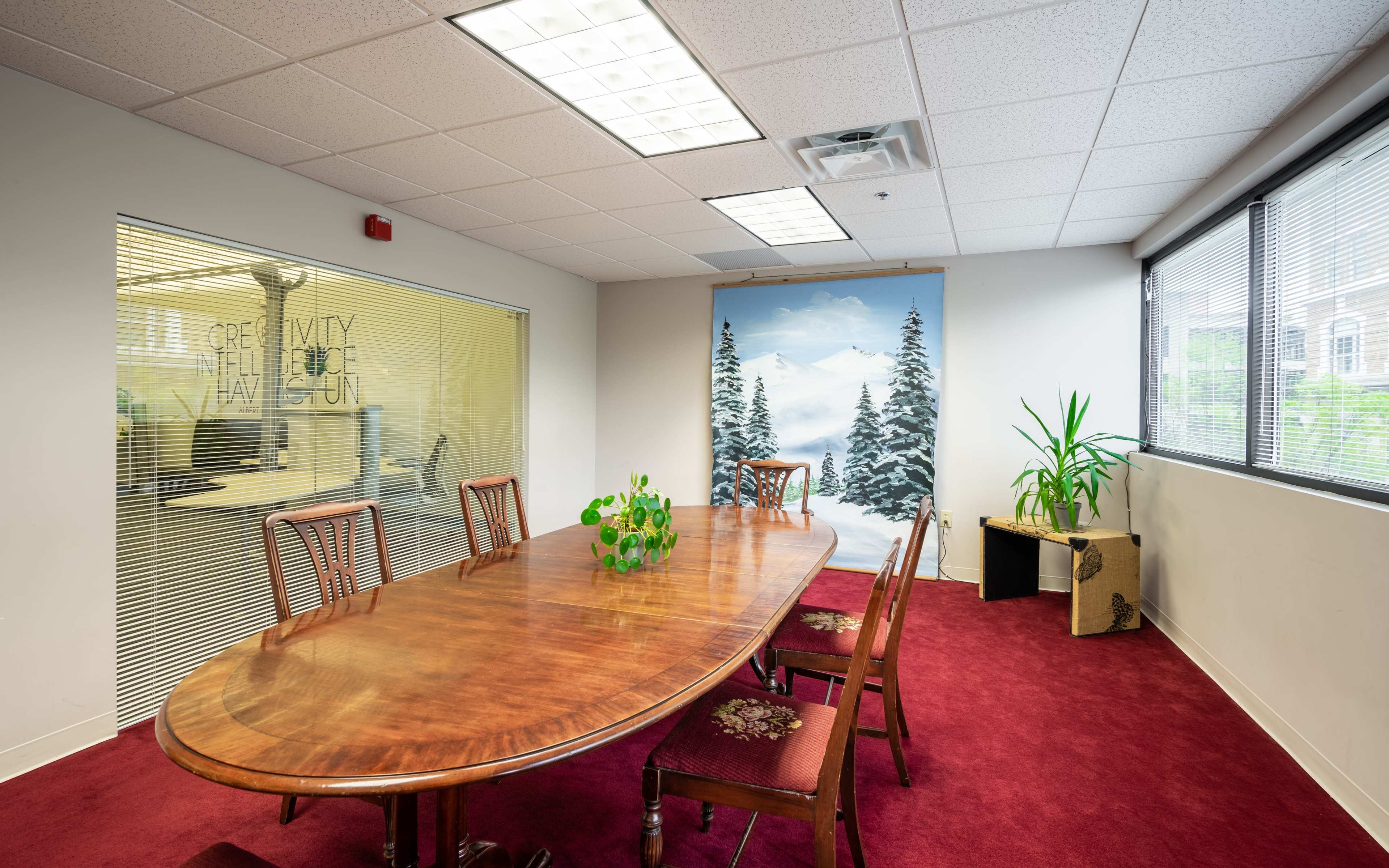A conference room features a large wooden table surrounded by chairs, with a snow-covered mountain mural on one wall and potted plants in the corner.