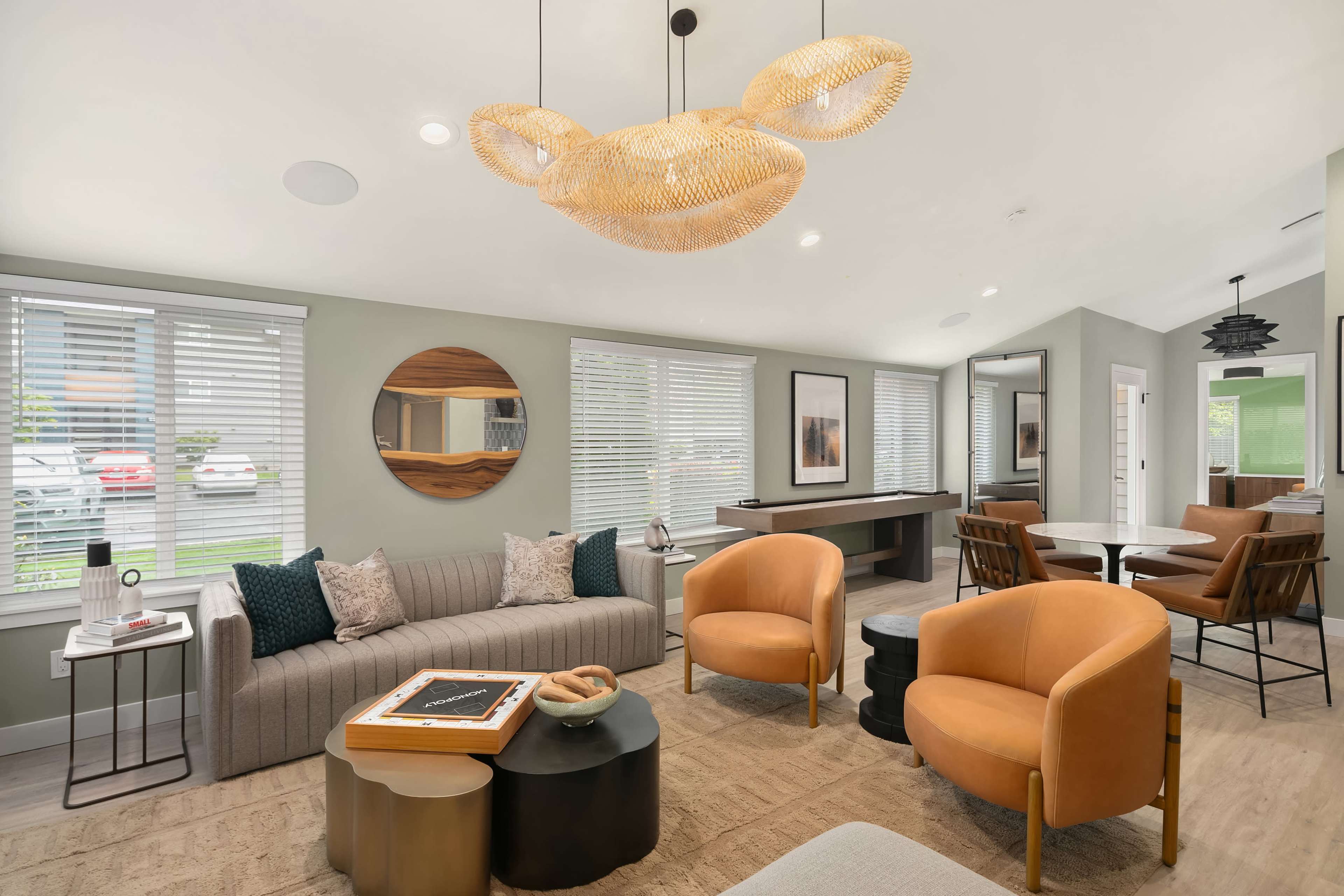 A modern living room features a mix of seating options, large windows with blinds, a round mirror, and a stylish light fixture.