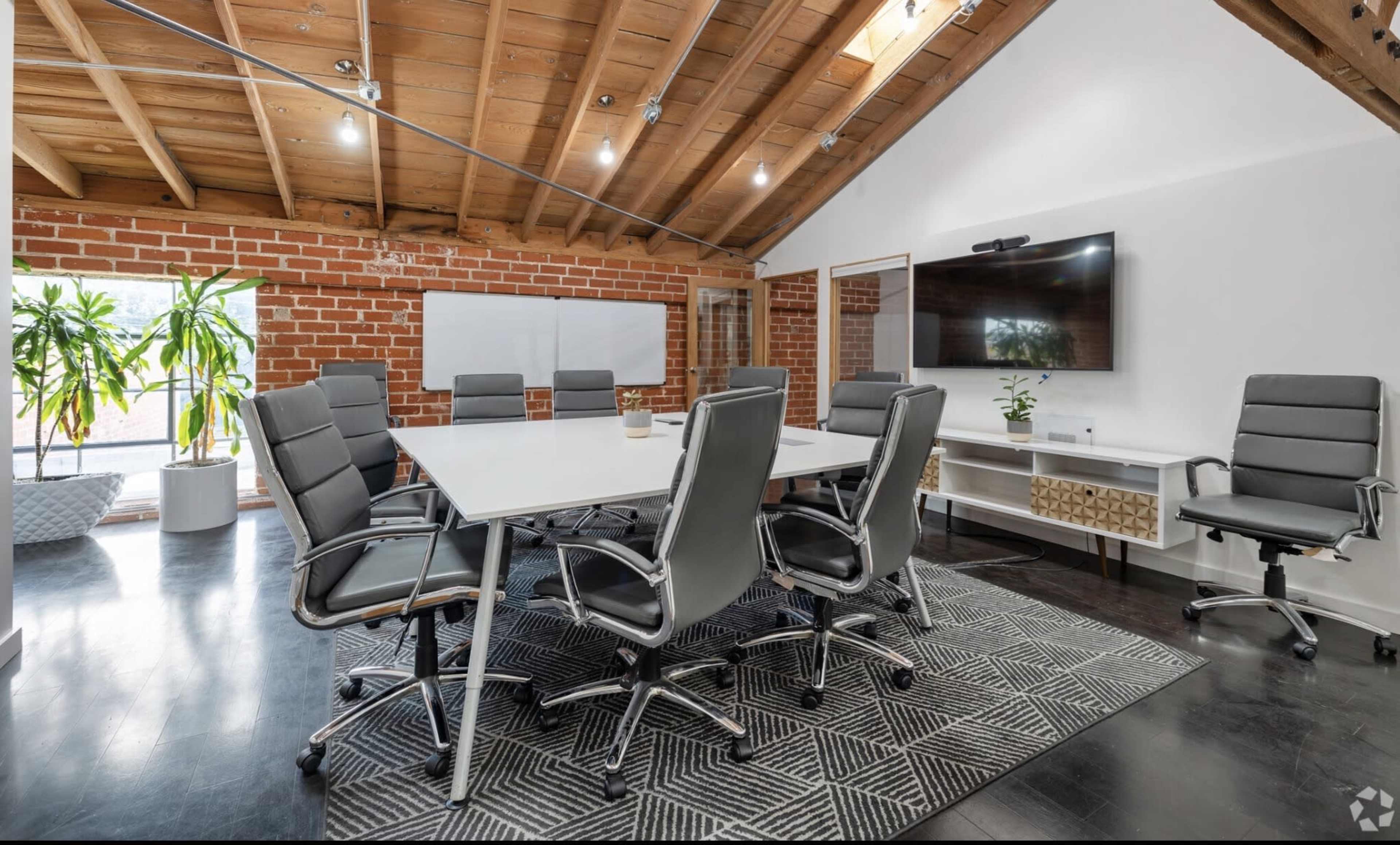 The image shows a modern conference room with a large rectangular table surrounded by gray ergonomic chairs, exposed wooden beams, and a wall-mounted television.