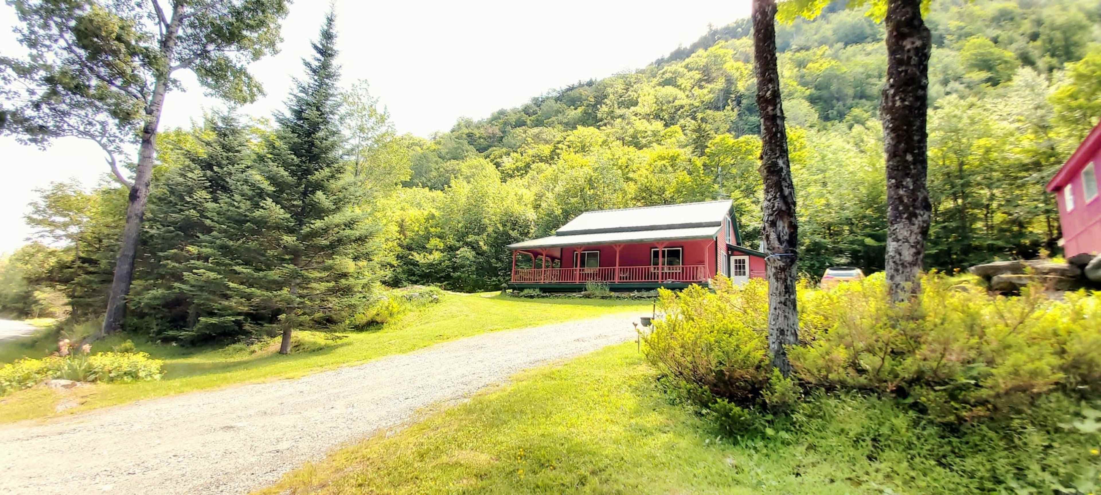 A red house with a porch is located beside a gravel driveway amidst green trees and a hillside.